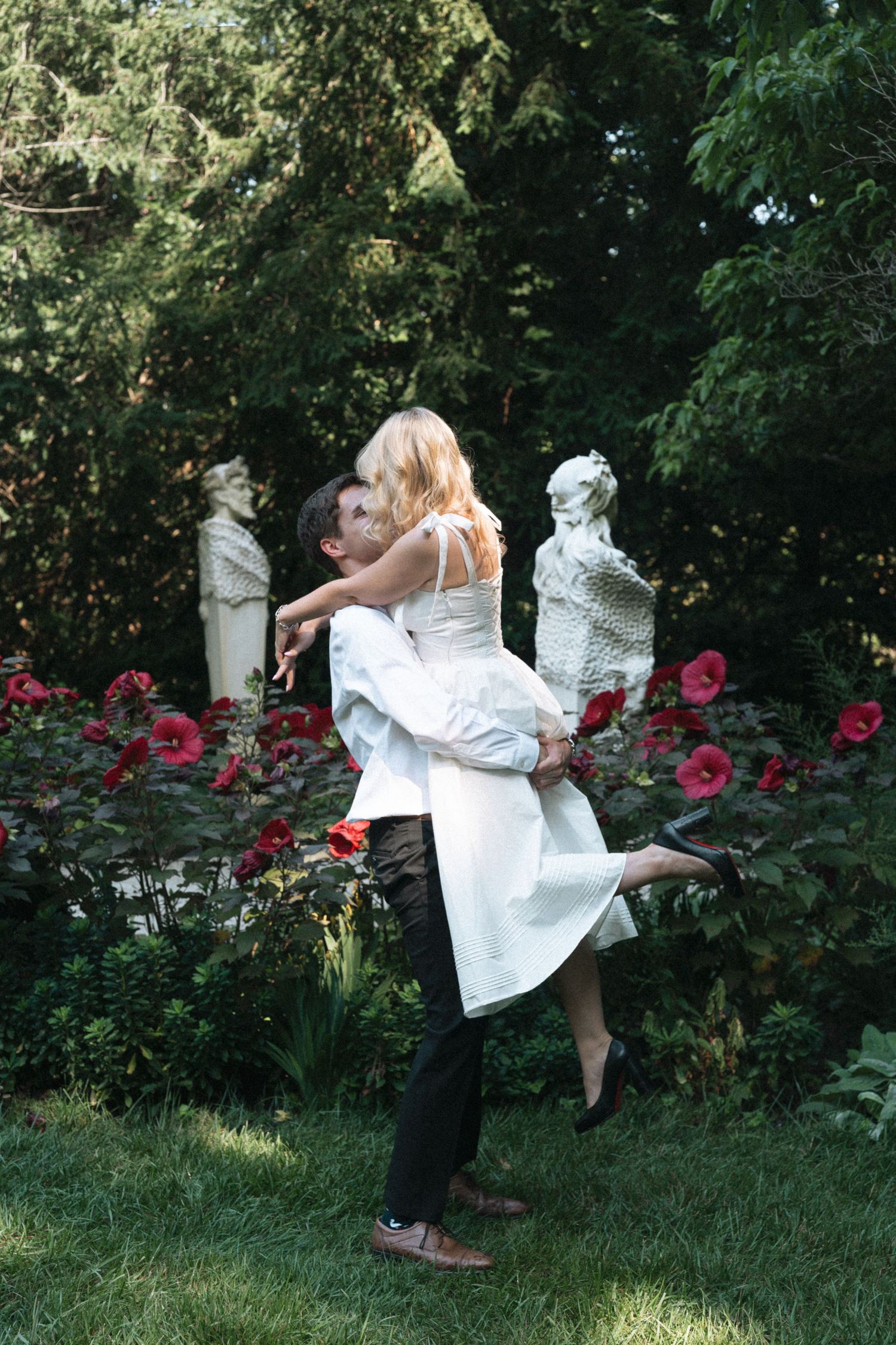 A couple embraces in a garden with red florals and decorative white statues, creating a romantic outdoor setting in Indianapolis