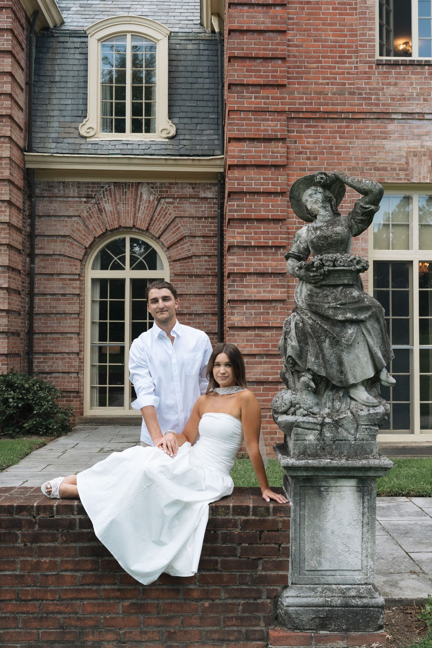 Elegant couple in white attire posing beside a stone statue outside a grand red brick mansion at the Kingwood Garden Center in Columbus