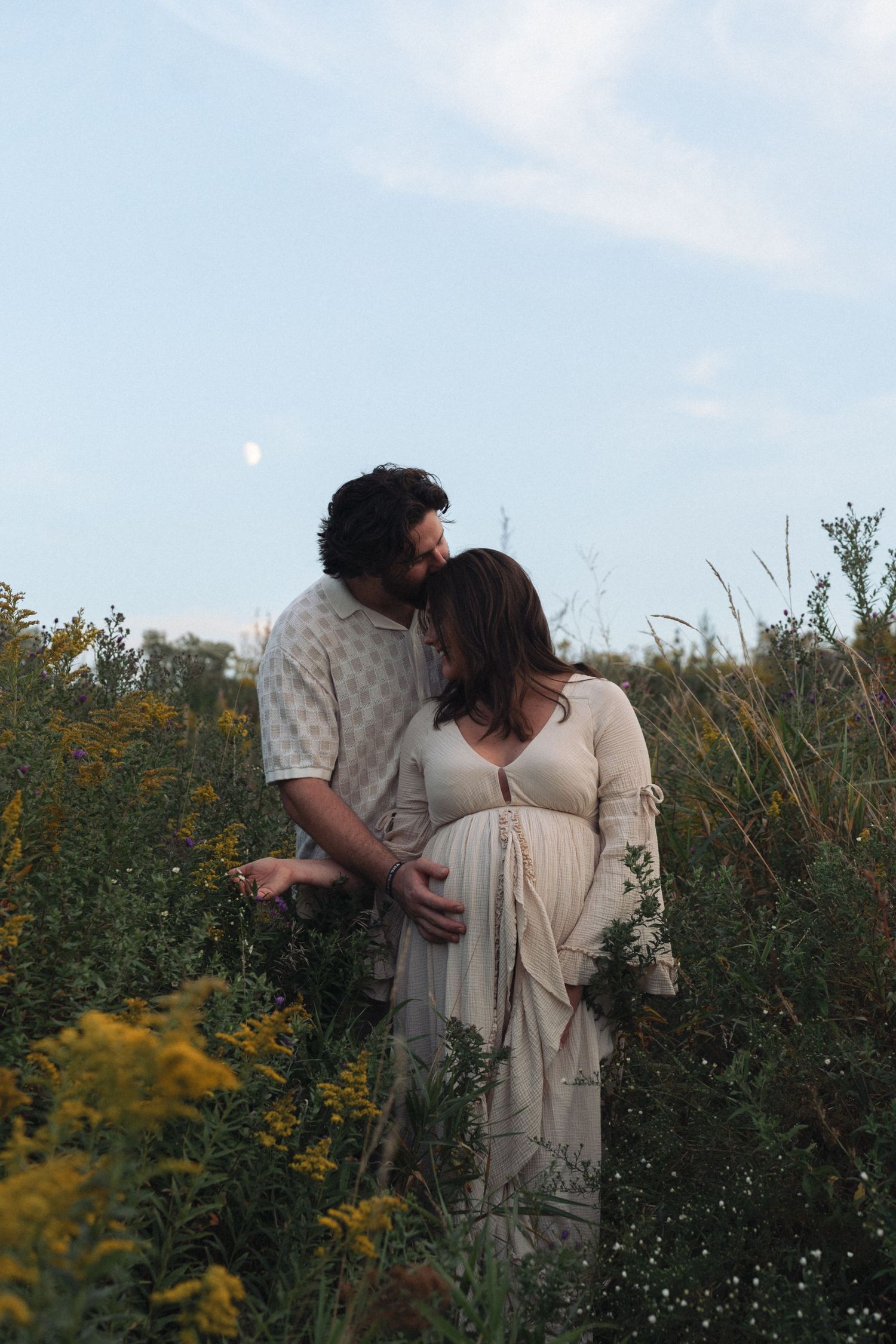 Couple embracing in a wildflower field at dusk, pregnant woman in white dress with partner kissing her head.