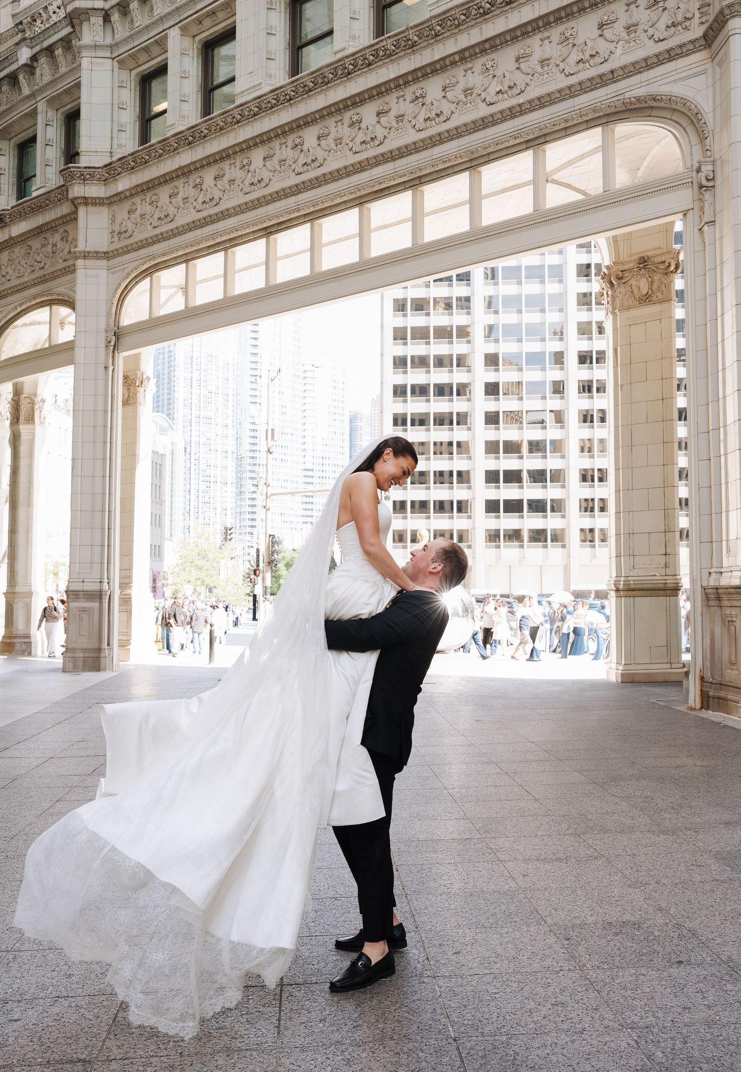 Groom lifts bride in white gown under ornate Wrigley Building arch, her veil flowing gracefully in a romantic wedding portrait in Chicago