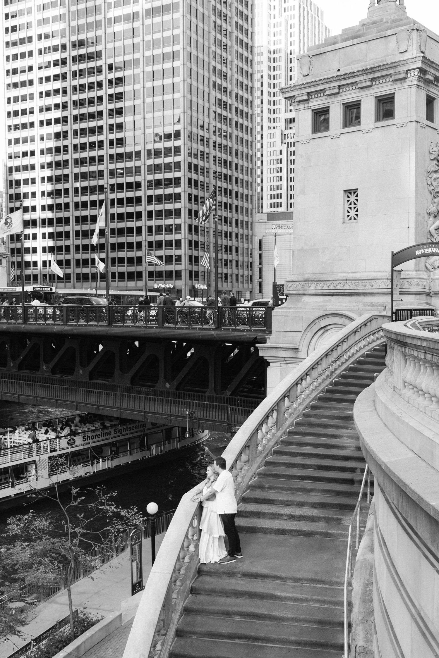 Black and white photo of a couple embracing on a curved stone staircase beside the Chicago River, with a bridge and skyscrapers behind them.