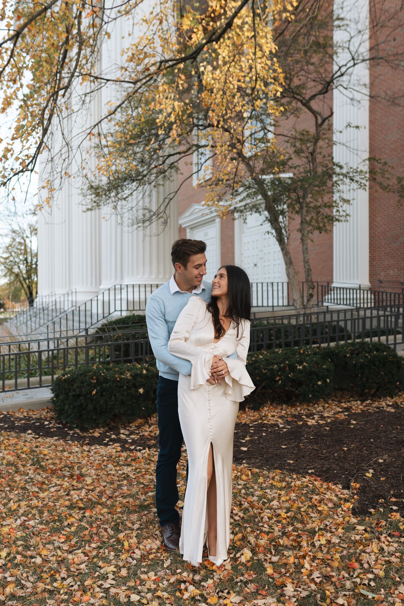 A romantic couple embraces on fallen autumn leaves in front of a columned building during golden hour in Wheaton, Illinois