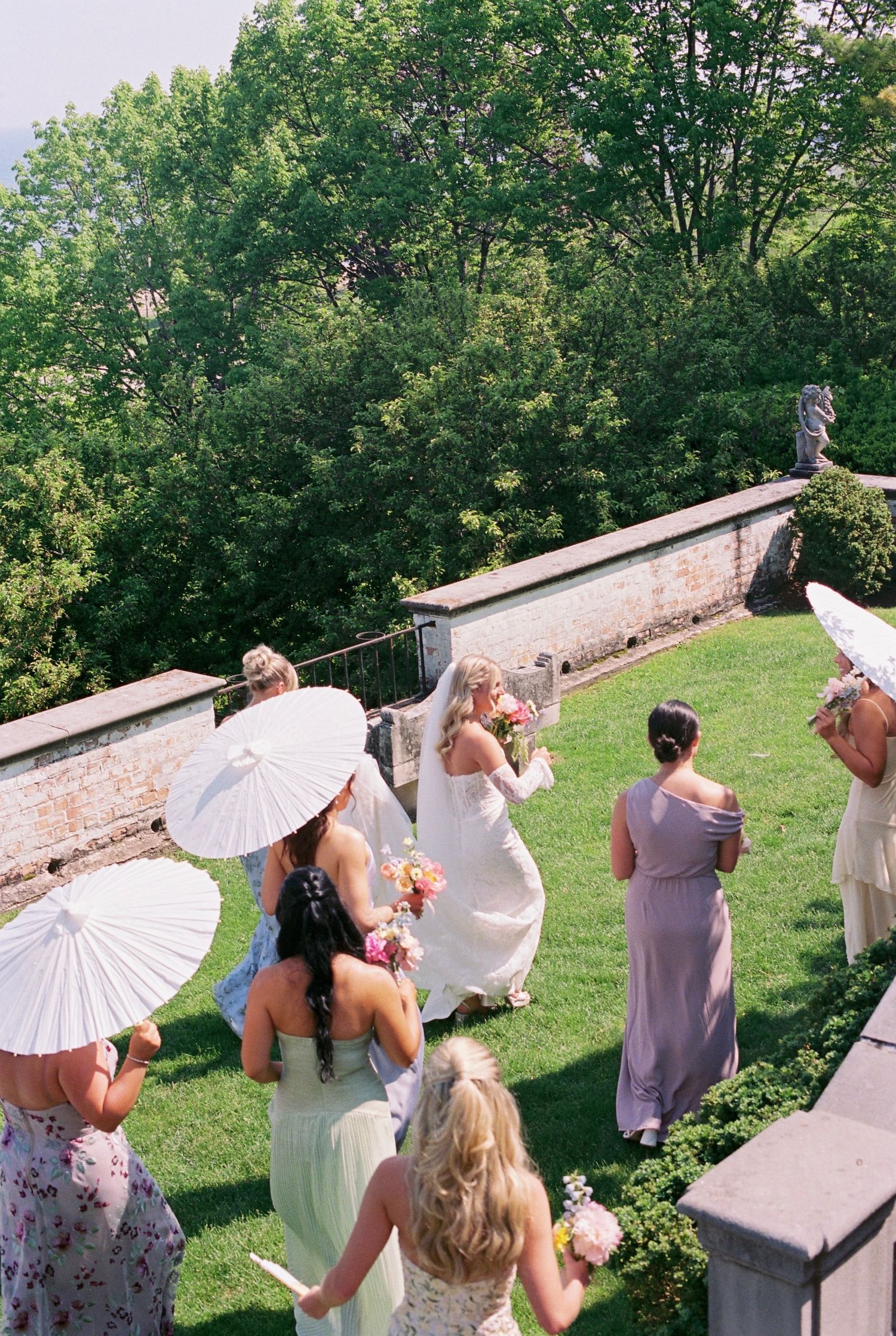 Wedding party holding white parasols gather on a grassy lawn near a stone wall during an outdoor summer celebration.