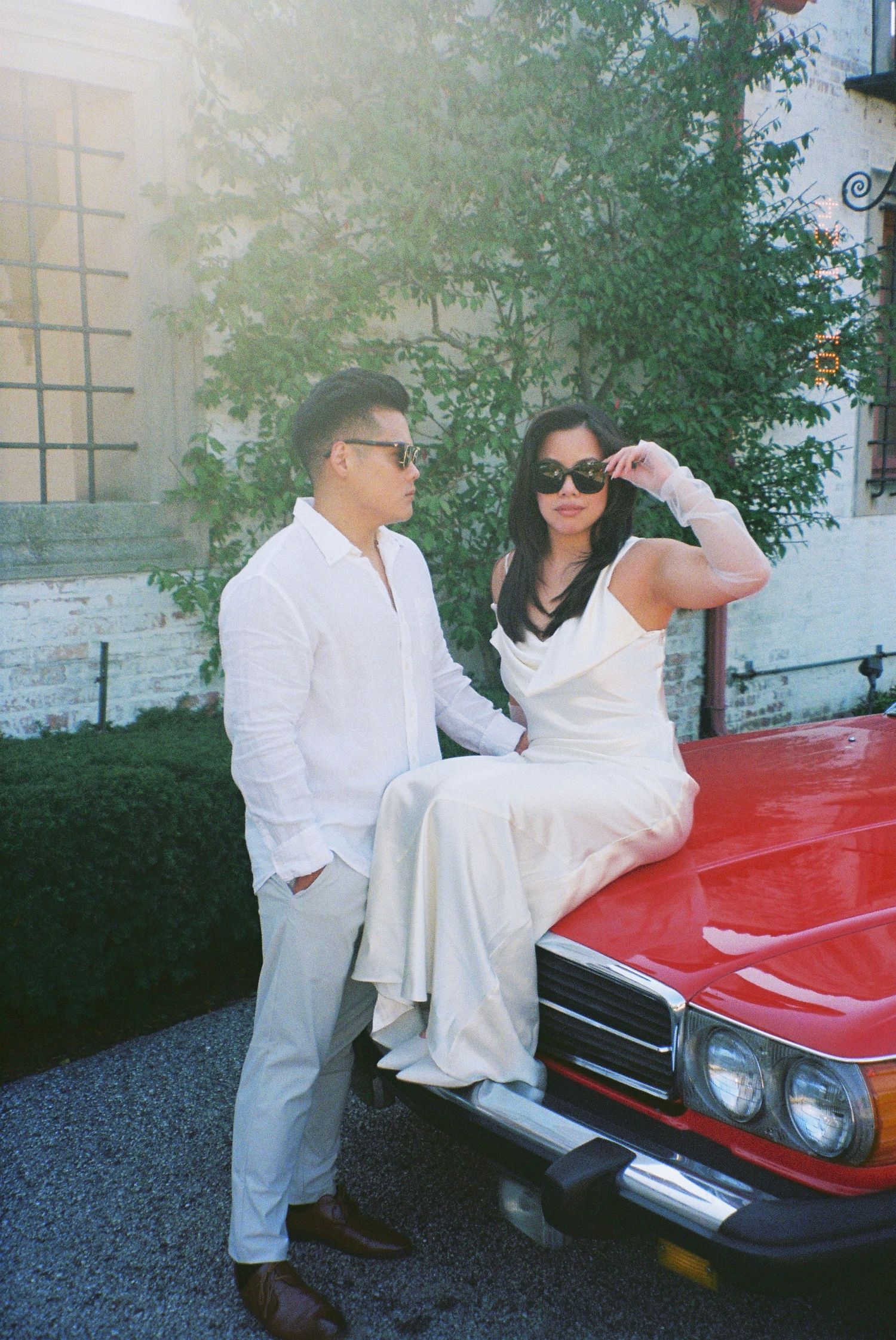 A person in white clothing and sunglasses leans against a vintage red car in a dappled sunlit driveway.