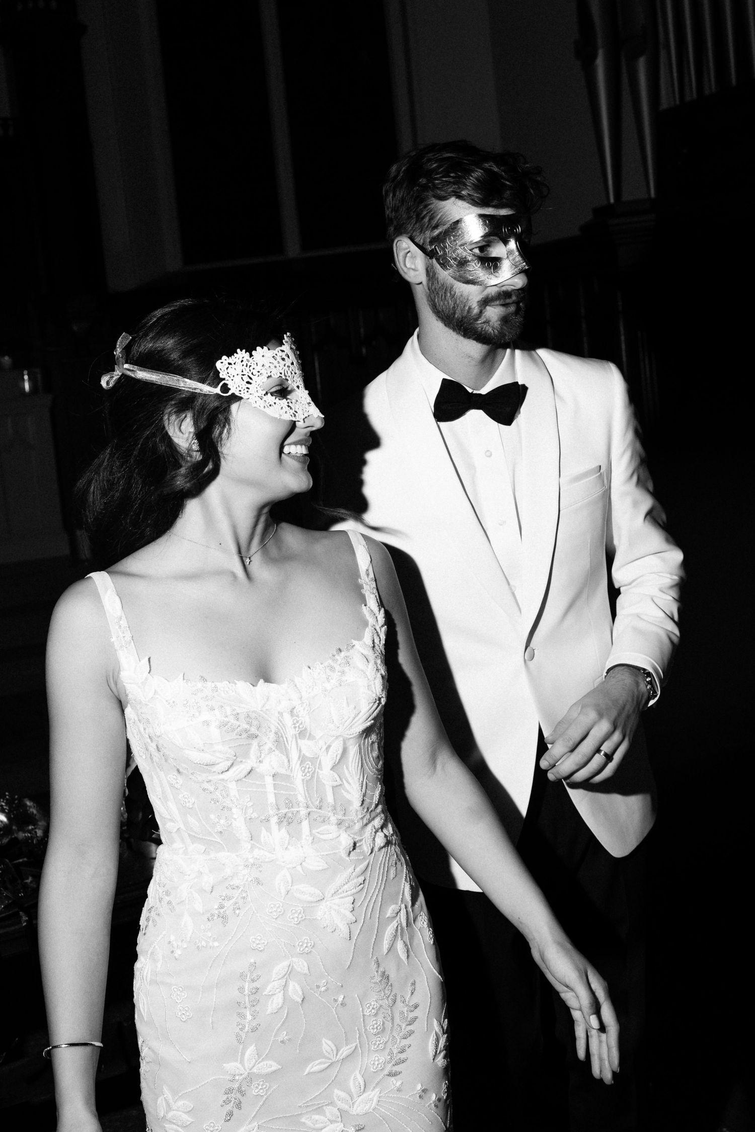 A bride in a white lace dress and groom in white tuxedo share candid laughing moments at their masquerade wedding reception