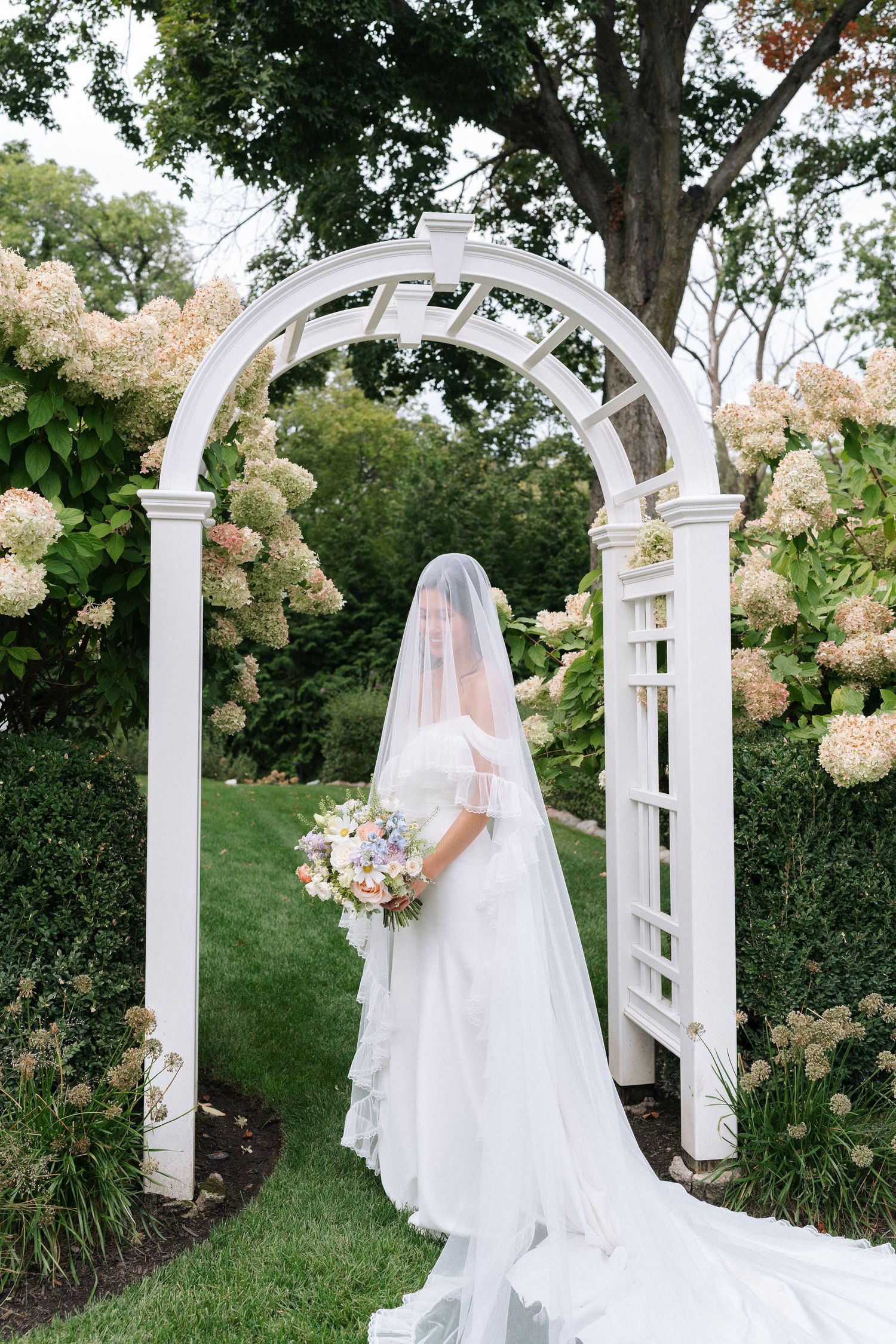 A New England bride draped in a delicate veil holding her bouquet against garden trellis arches adorned with hydrangeas