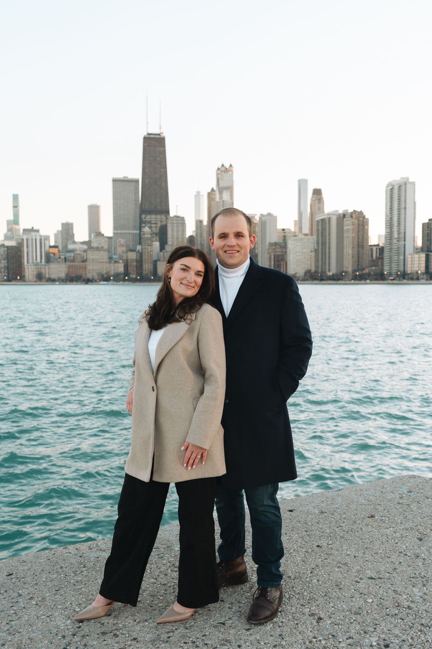 A couple poses together on a stone breakwall with the Chicago skyline and Lake Michigan creating a scenic backdrop.