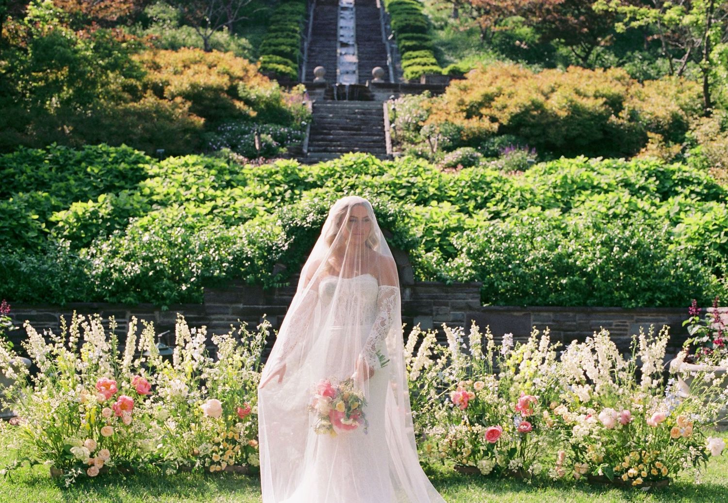 wedding dress gently sways in a lush garden setting with blooming flowers and greenery.