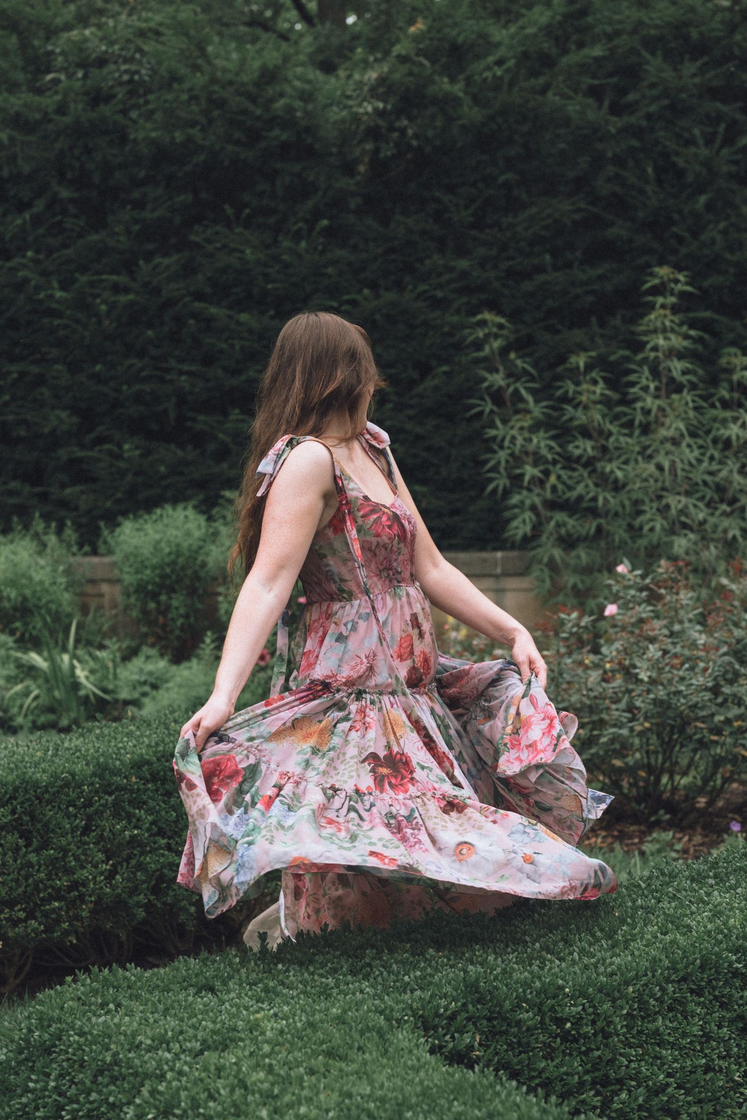 A girl in floral dress sits gracefully on green grass near garden hedges in a dreamy outdoor setting at Newfields in Indianapolis