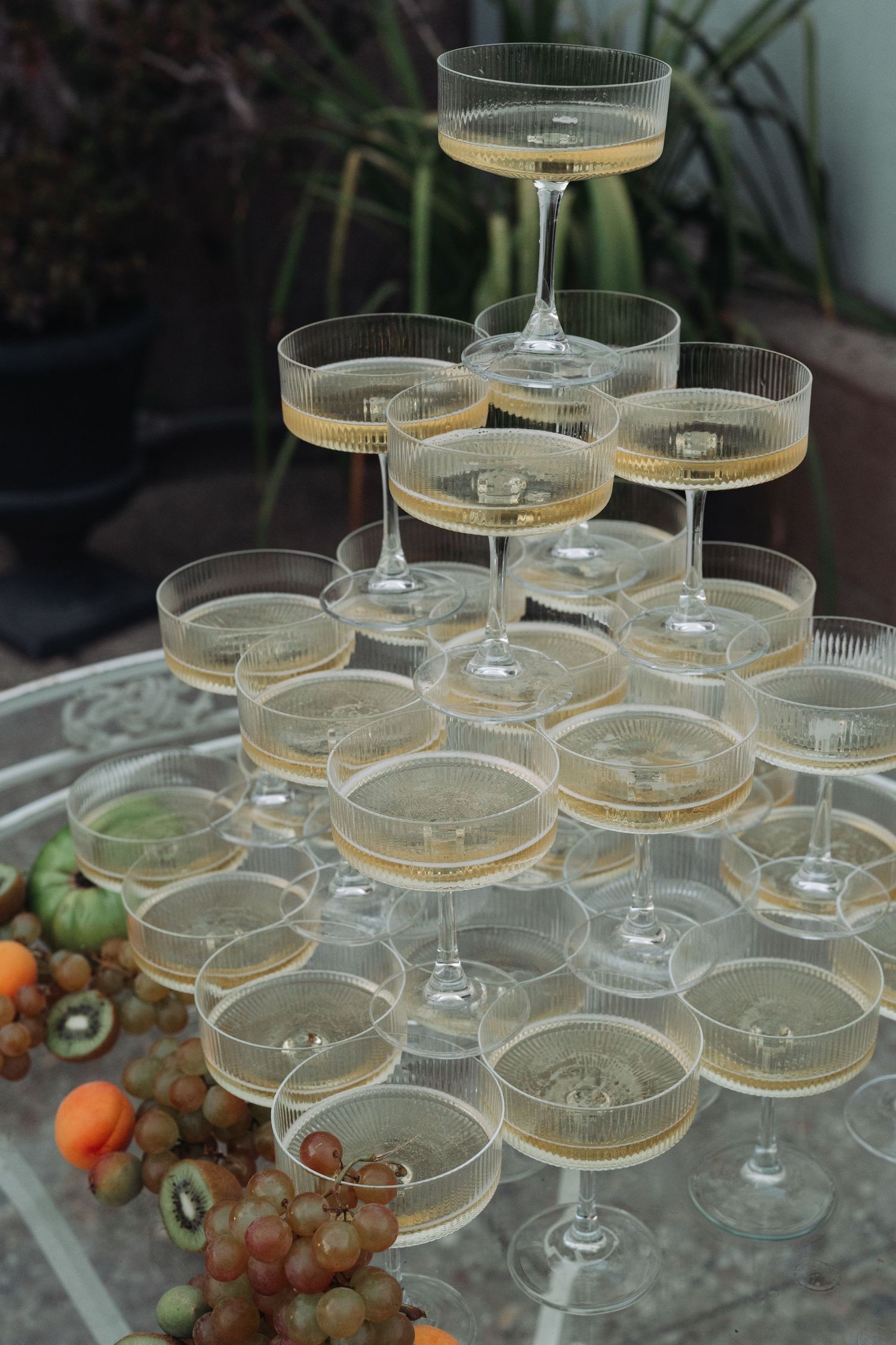 A champagne tower with stacked coupe glasses arranged in tiers next to fresh fruits and grapes on a table.