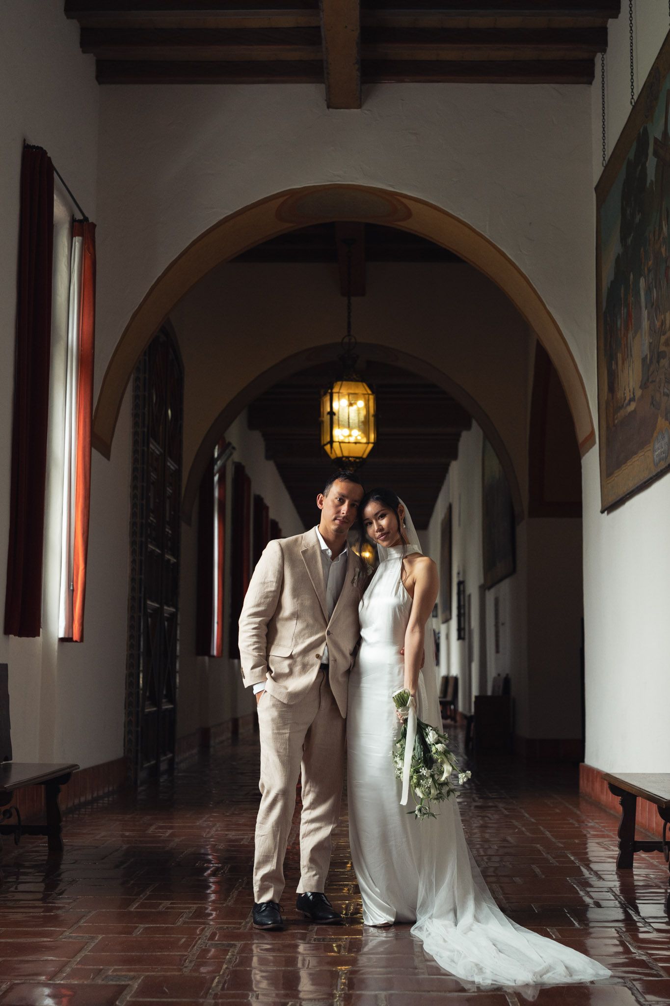 A couple poses in an elegant Spanish colonial hallway with arches and terra cotta floors for their wedding portrait in Santa Barbara