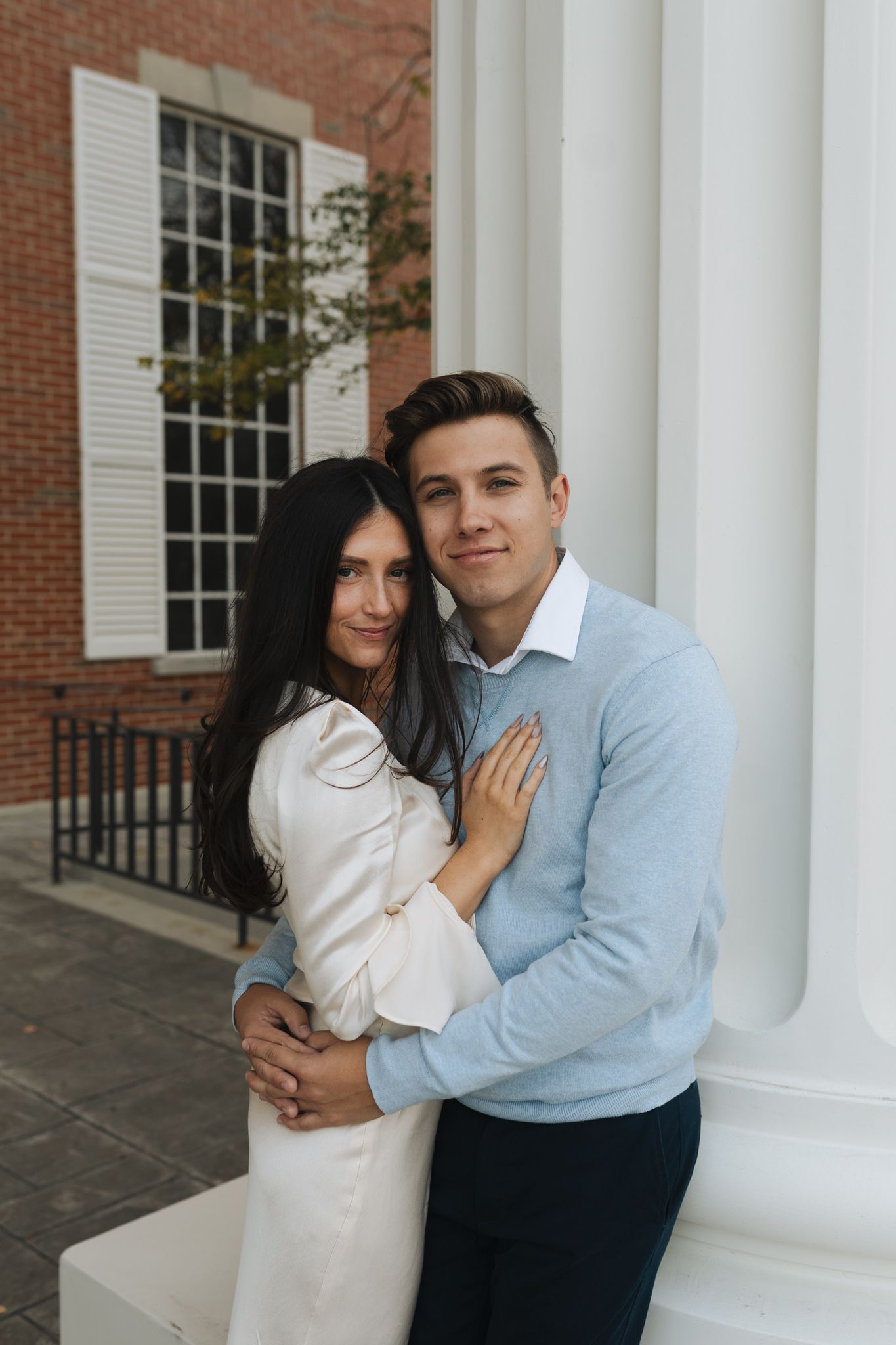 A couple poses together for romantic engagement photos against white columns and brick walls of a historic building at Wheaton College