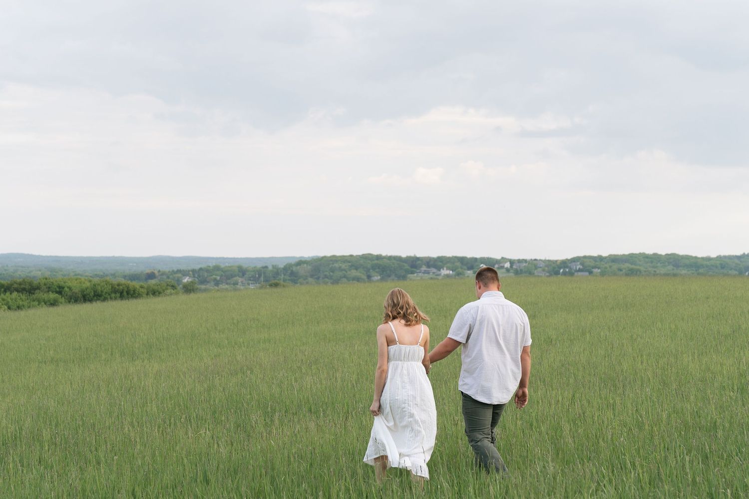 Couple holding hands walking through a lush green field with rolling hills and cloudy sky in background.