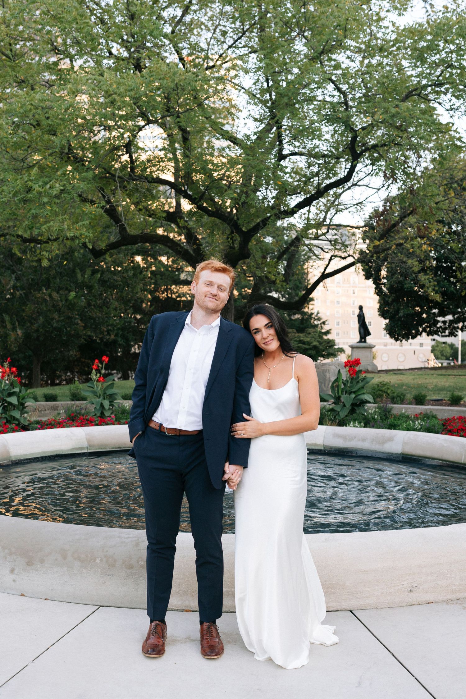 A couple poses together on a curved stone bench in a garden setting with trees and red flowers at the Tennessee State Capitol