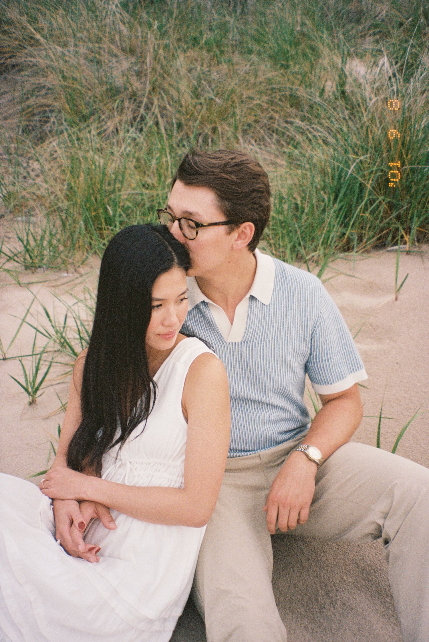 A couple in white and blue outfits share tender moments while sitting on a sandy beach with grass in the background.