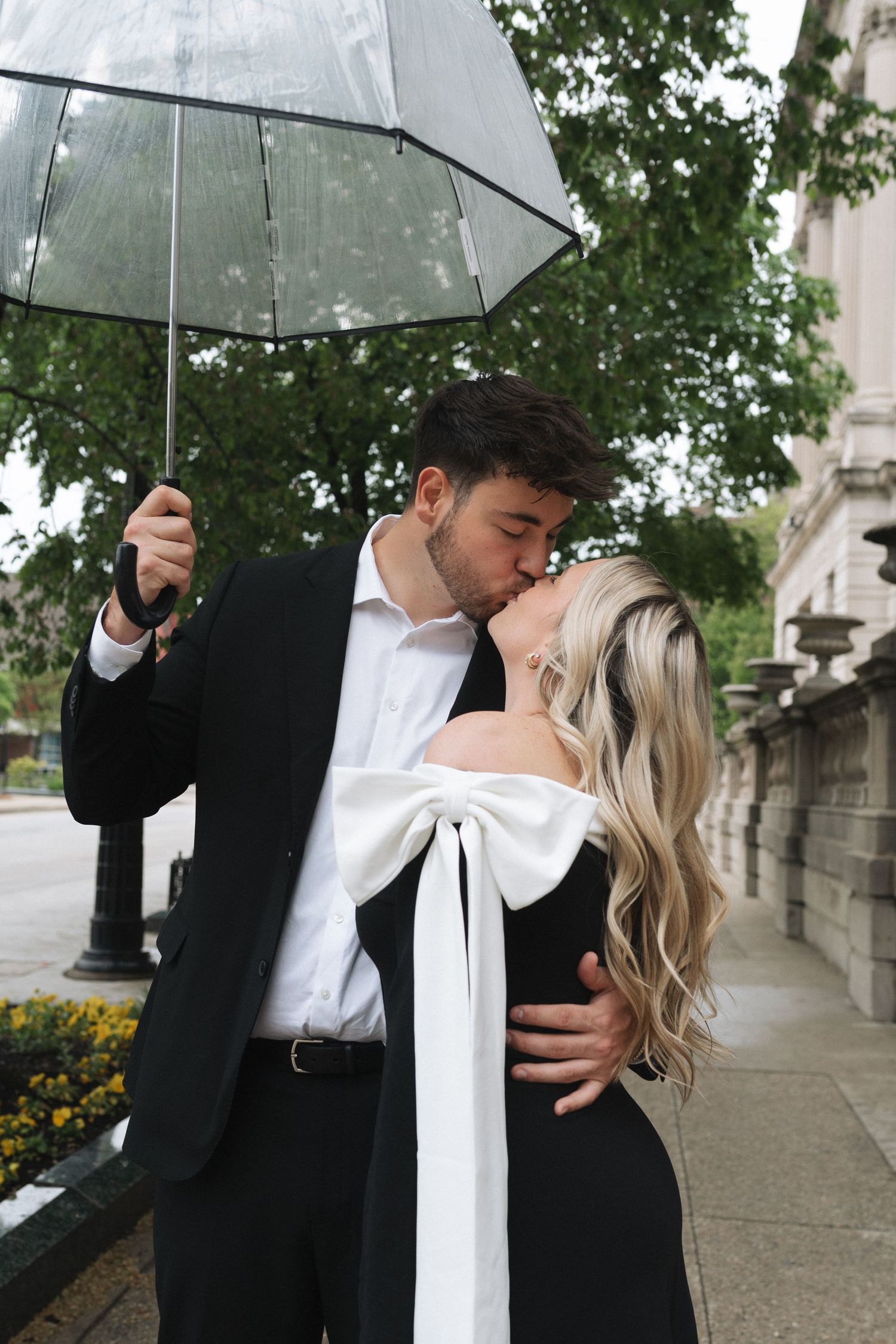 A couple shares romantic moments under a clear umbrella on a rainy day, dressed in formal black and white attire.