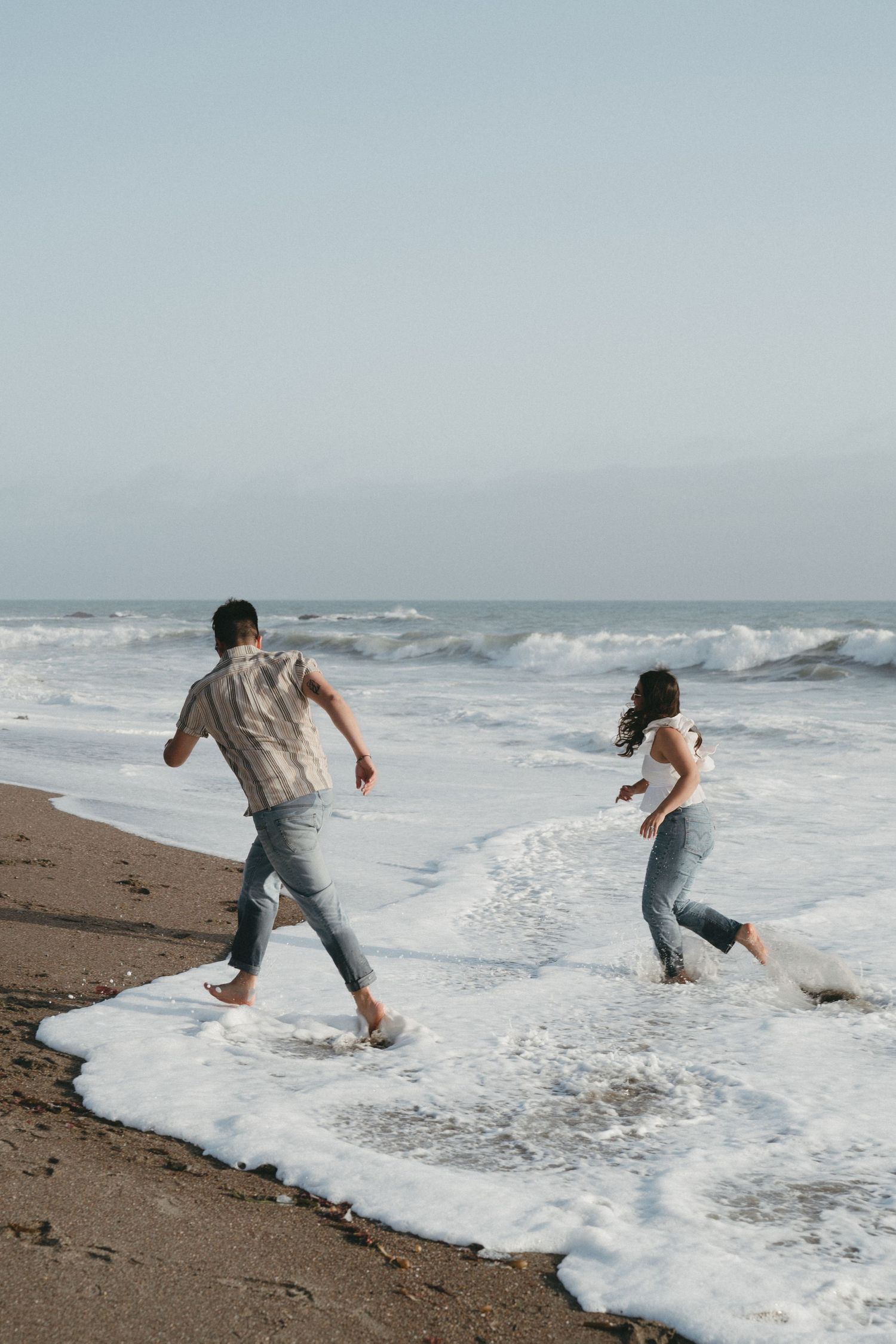 A couple playfully running and jumping through ocean waves breaking on a sandy beach in Cambria, California