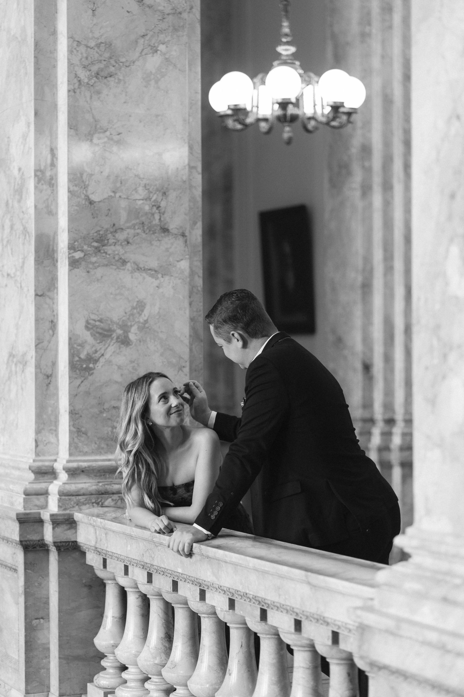 Black and white photo of an elegant couple sharing a romantic moment on a marble balcony with chandelier.