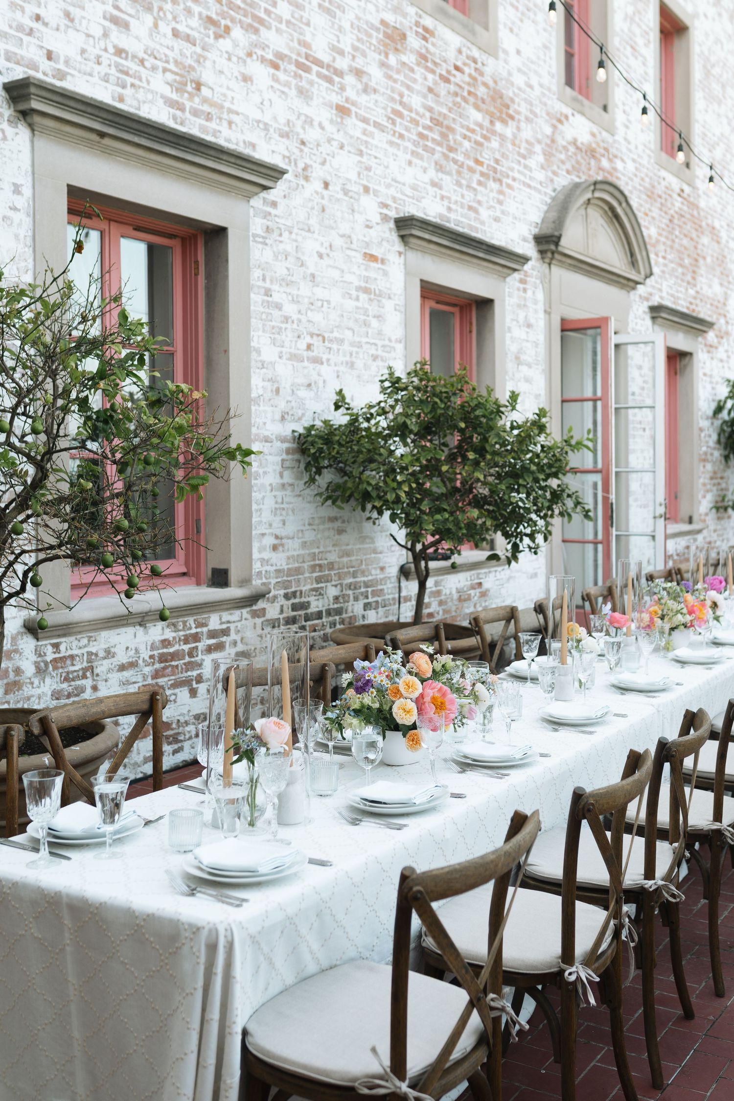 A rustic wedding reception table setting with white linens, dark wood chairs, and potted greenery along a brick wall.