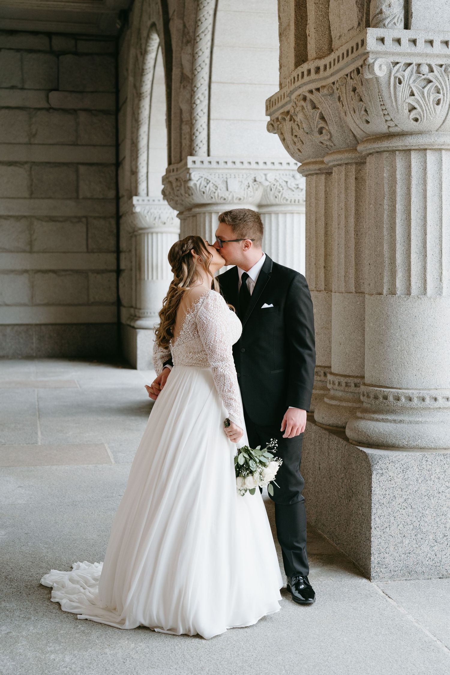 Bride and groom sharing a kiss beside ornate stone columns in a grand architectural setting.