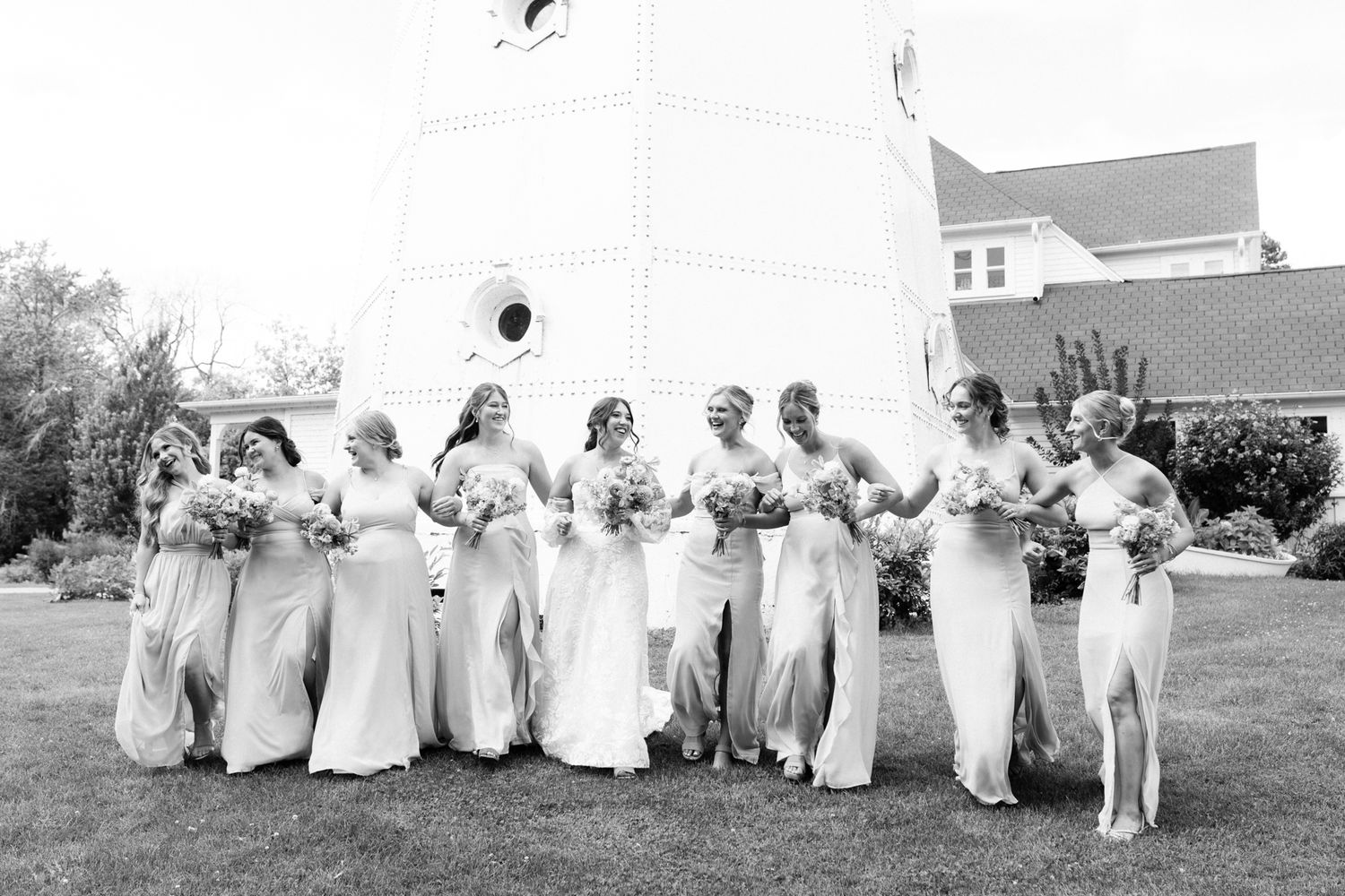 Black and white photo of bride and bridesmaids in long gowns holding bouquets outdoors near a white lighthouse