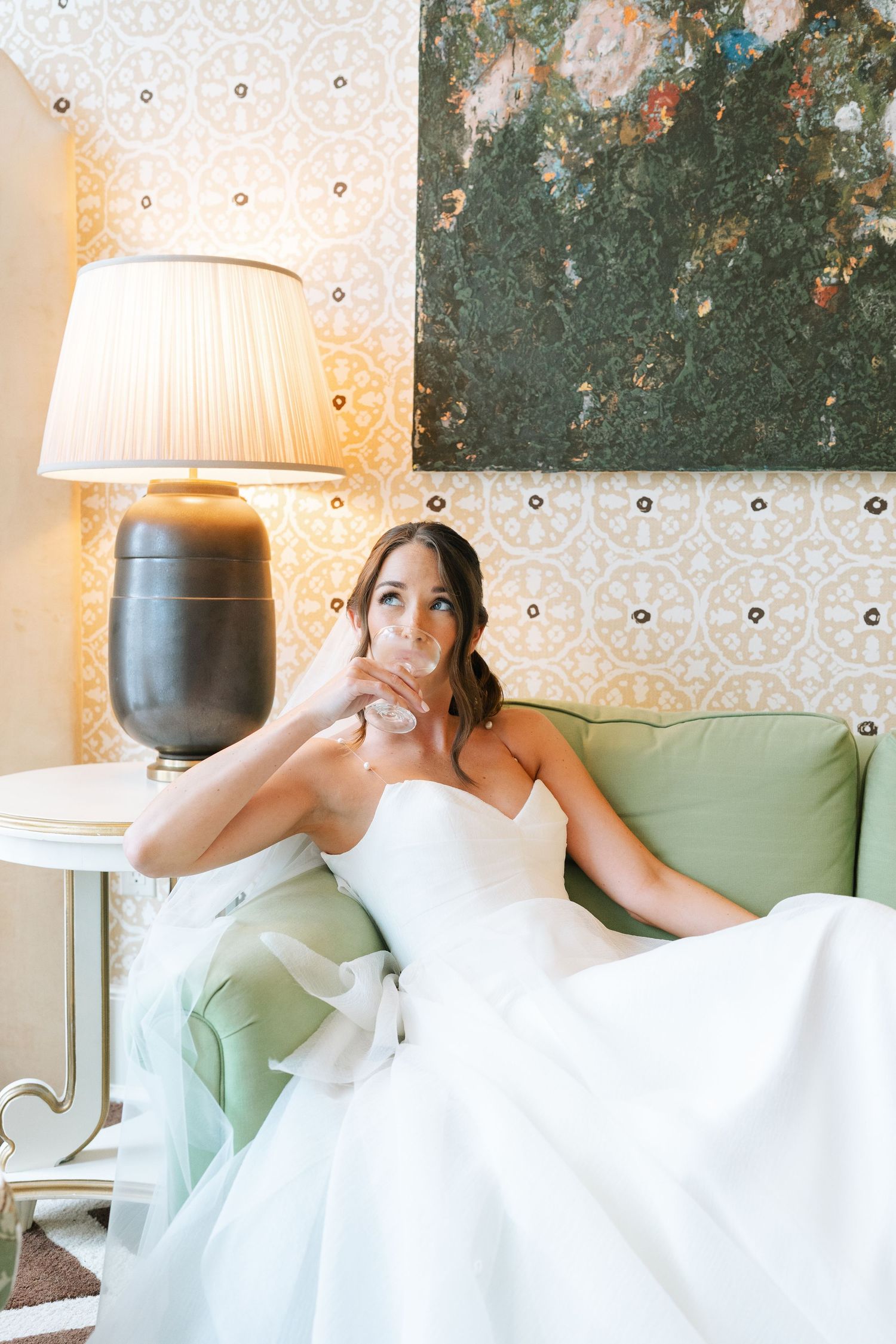 Luxury bride reclining on mint green sofa with decorative wallpaper and table lamp in vintage-style setting sipping champagne