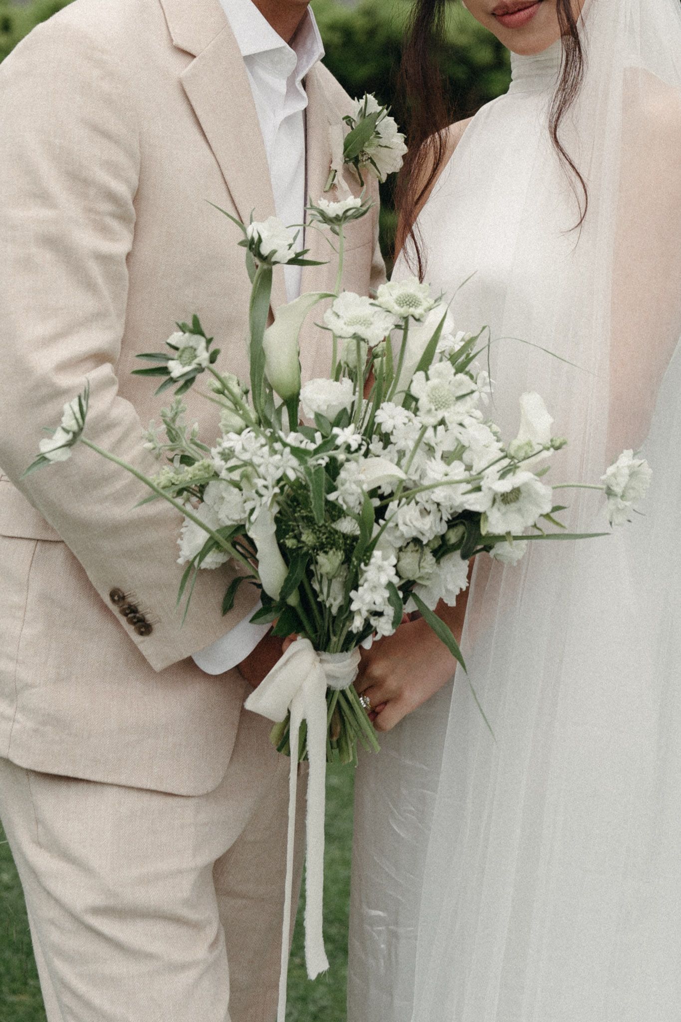 Close-up of wedding bouquet with white flowers and ribbons against tan suits and white dress details in outdoor setting in California