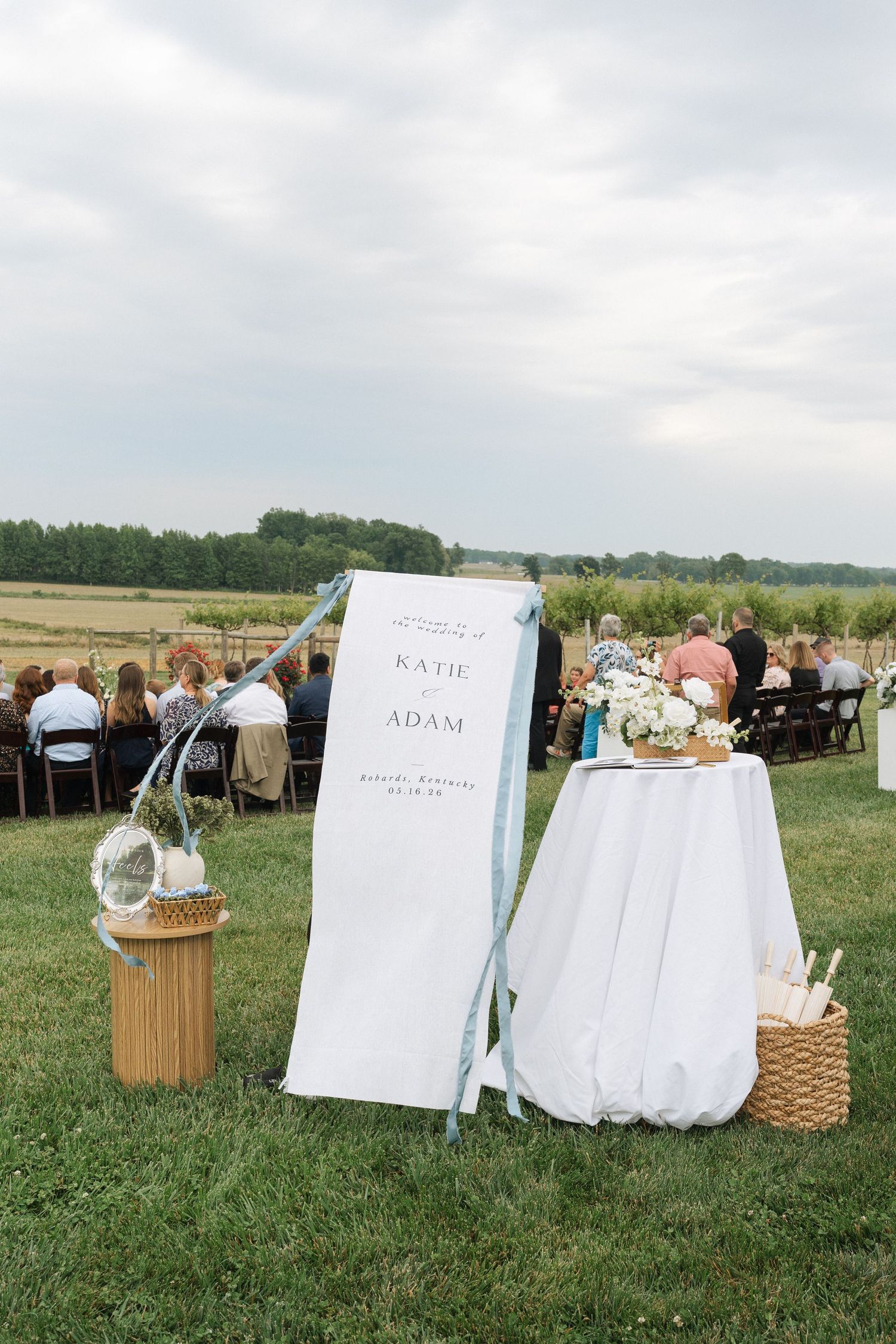 Outdoor wedding ceremony setup with a Katie and Adam welcome banner, floral table and guests seated on a grassy field in Kentucky