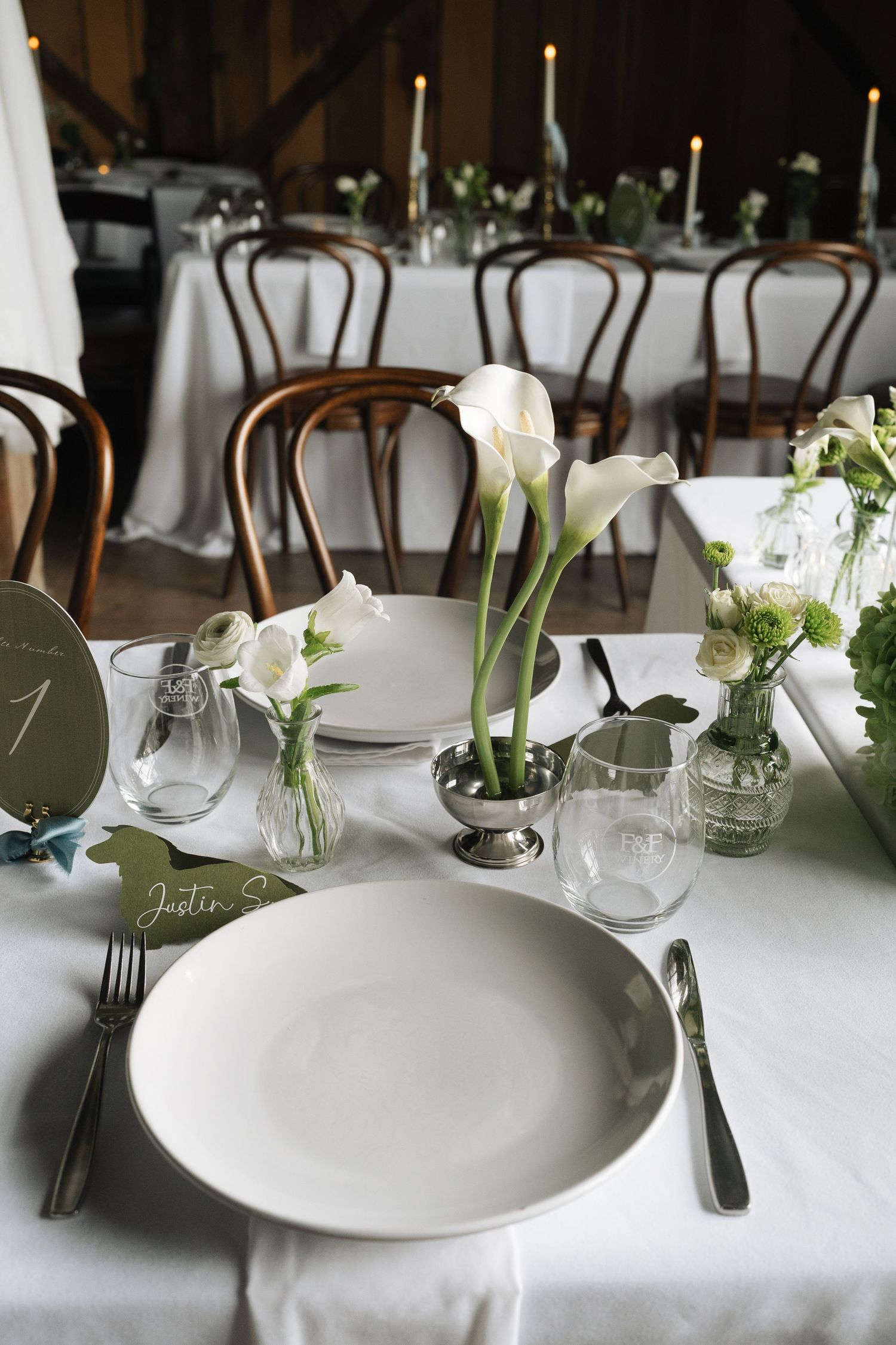 Elegant wedding table setting with white plates, calla lily centerpieces, glassware, and a green place card, with candlelit tables behind