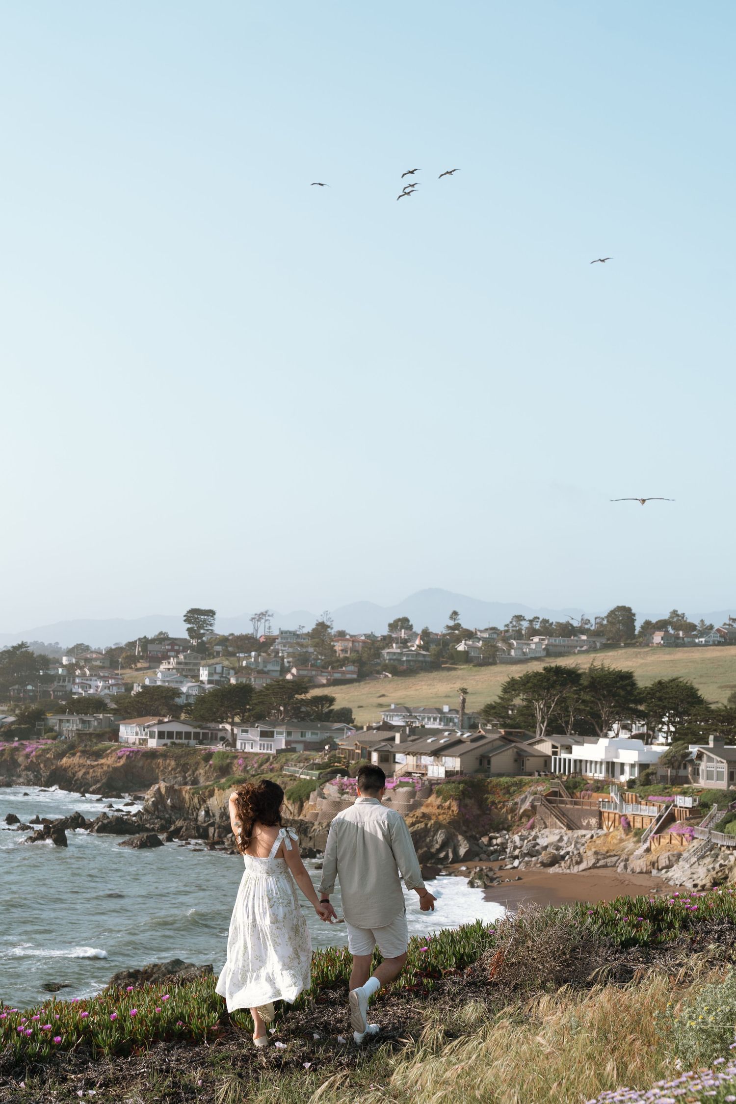 Two people in white outfits stand at a coastal overlook enjoying views of a California beach town and rocky shoreline.