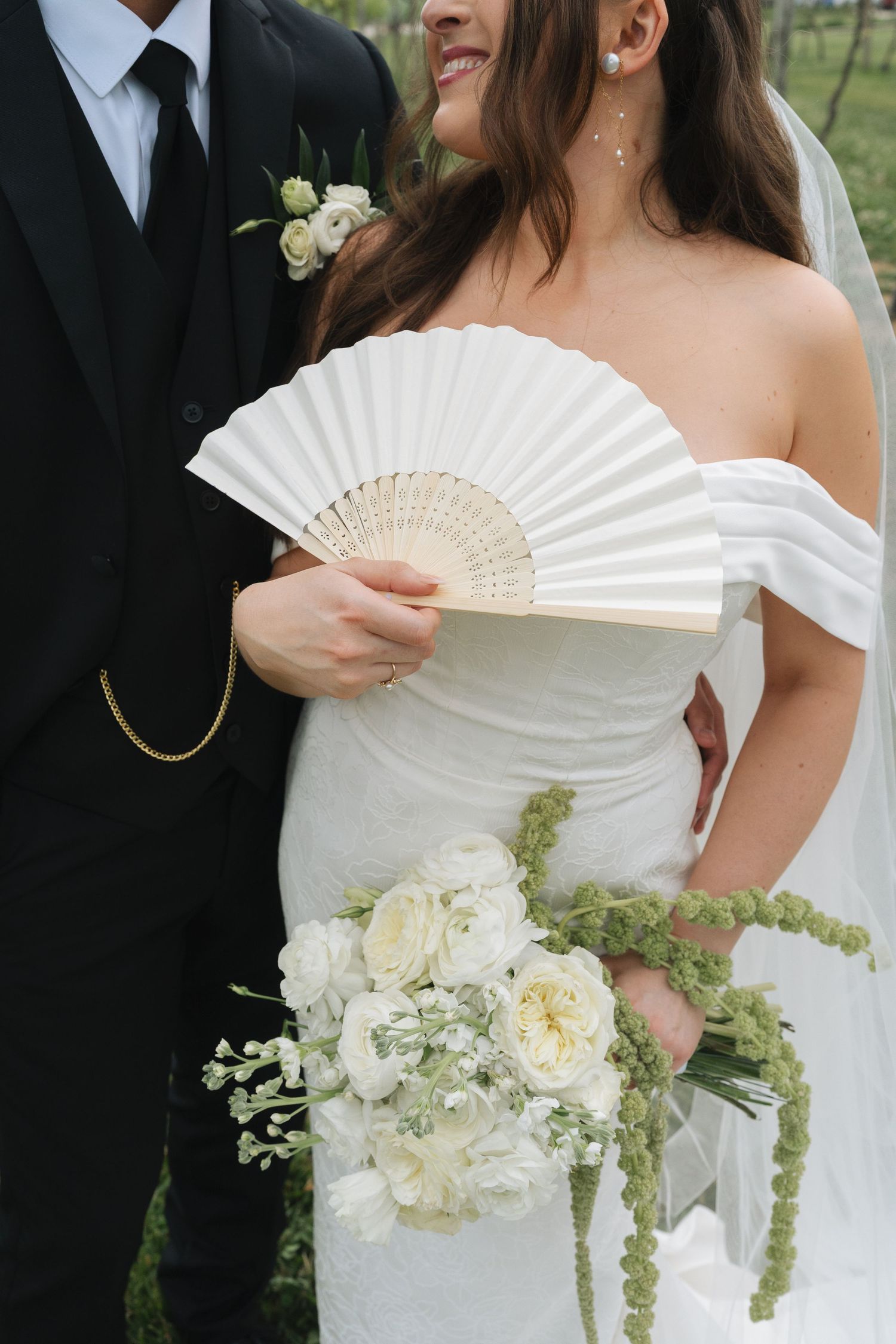 Bride in off-shoulder white gown holds floral bouquet and white hand fan beside groom in black suit in Kentucky