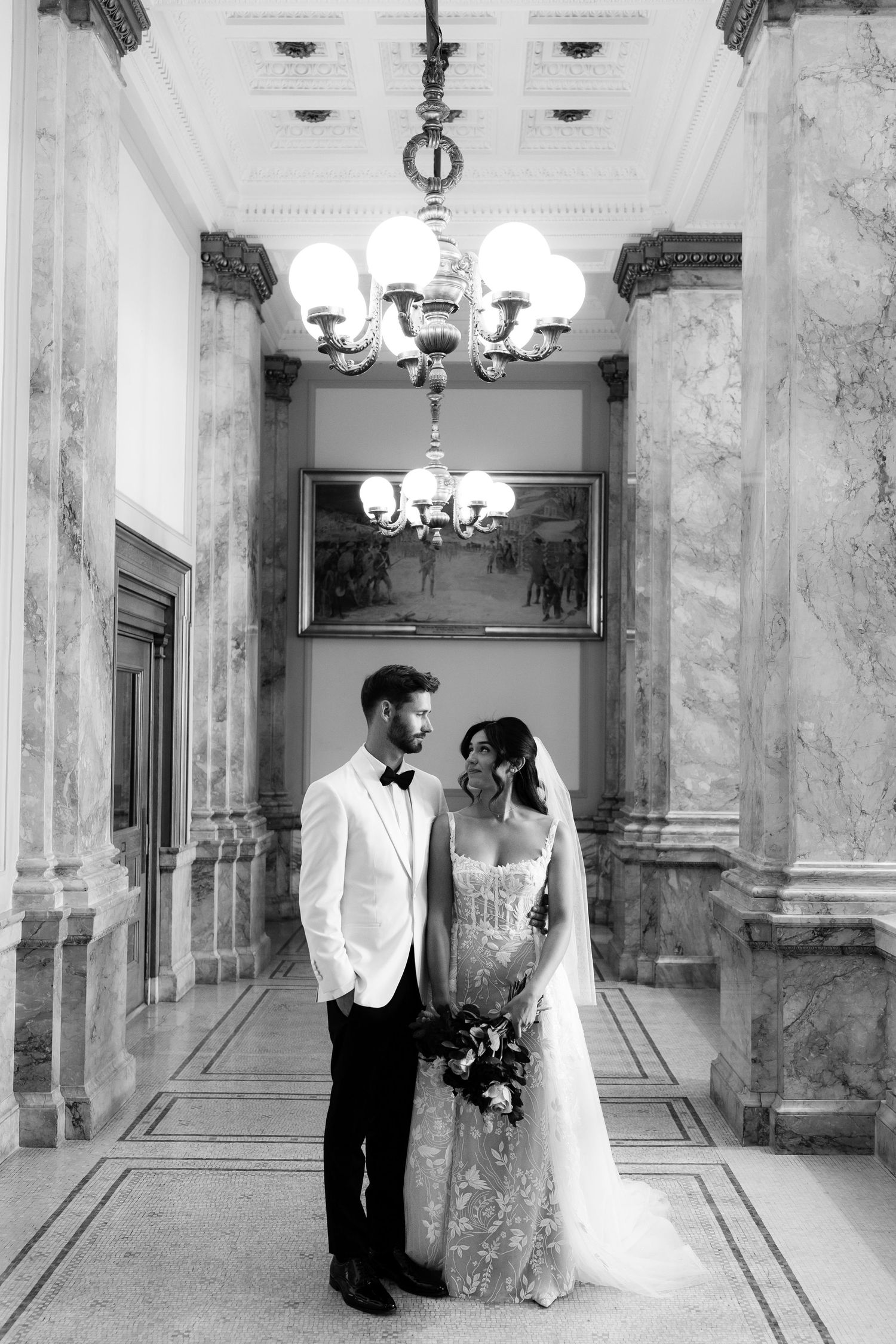 Black and white wedding portrait of bride and groom in elegant marble hall with chandelier.