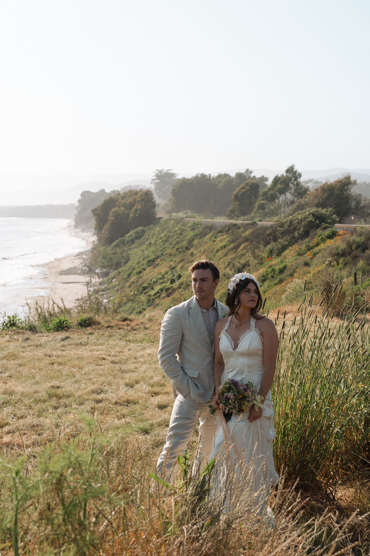 Romantic couple pose together along scenic coastal cliffside with tall grass and ocean views at sunset in California
