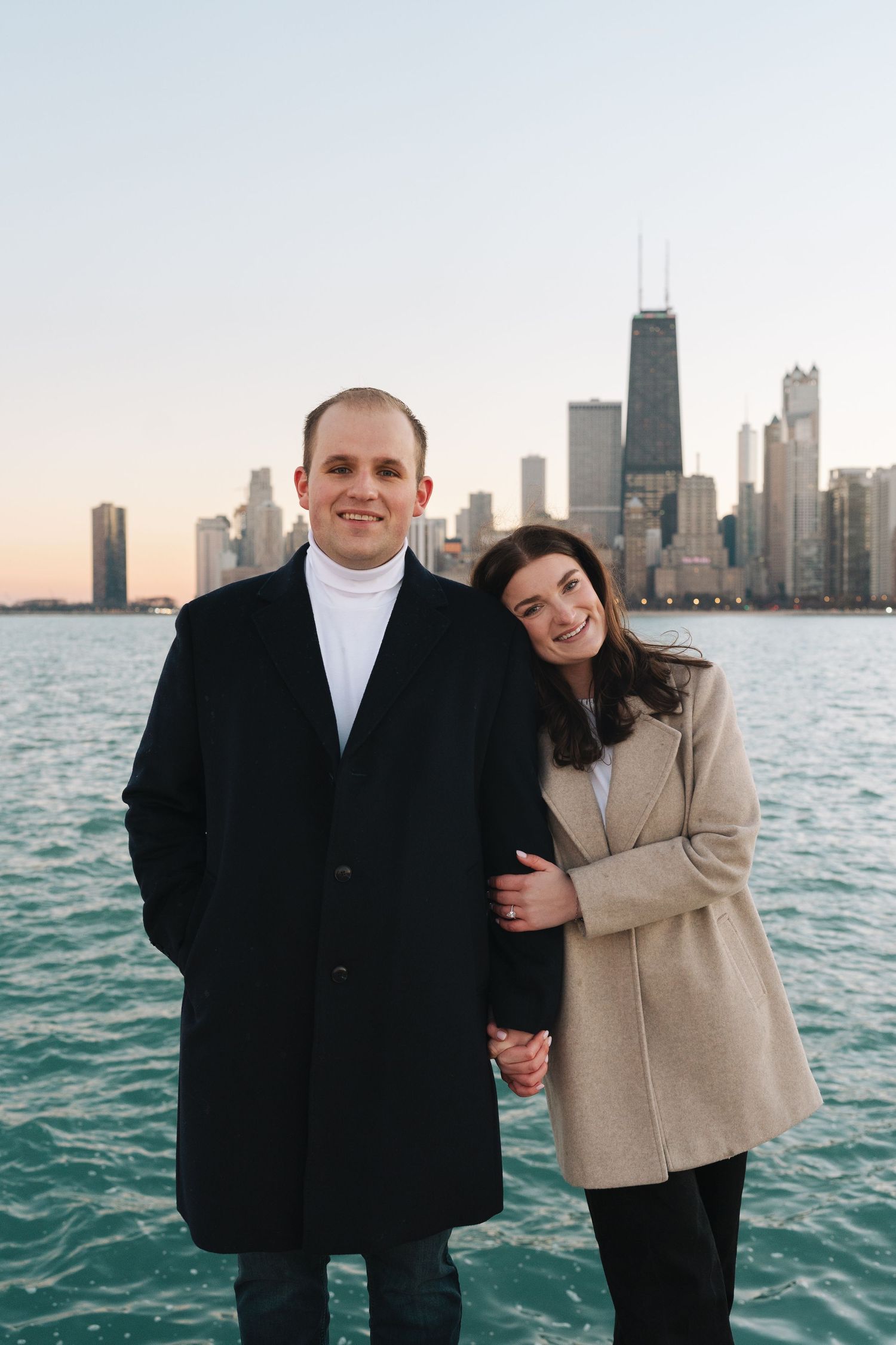 A couple stands along Lake Michigan with the Chicago skyline in the background during sunset.
