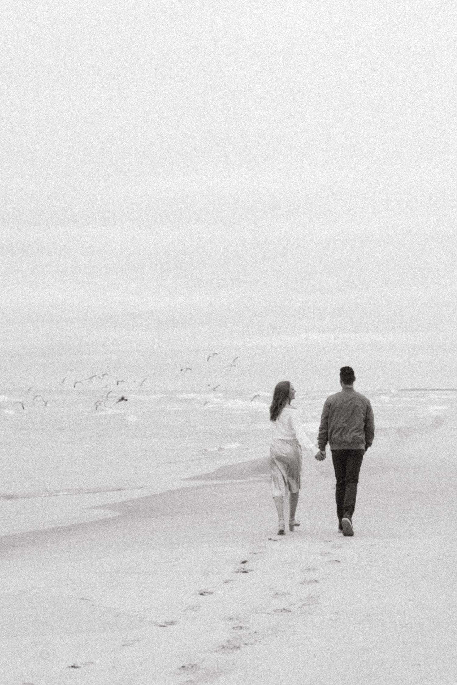 Black and white photo of a couple holding hands walking on an ocean beach with seagulls flying in the misty background.