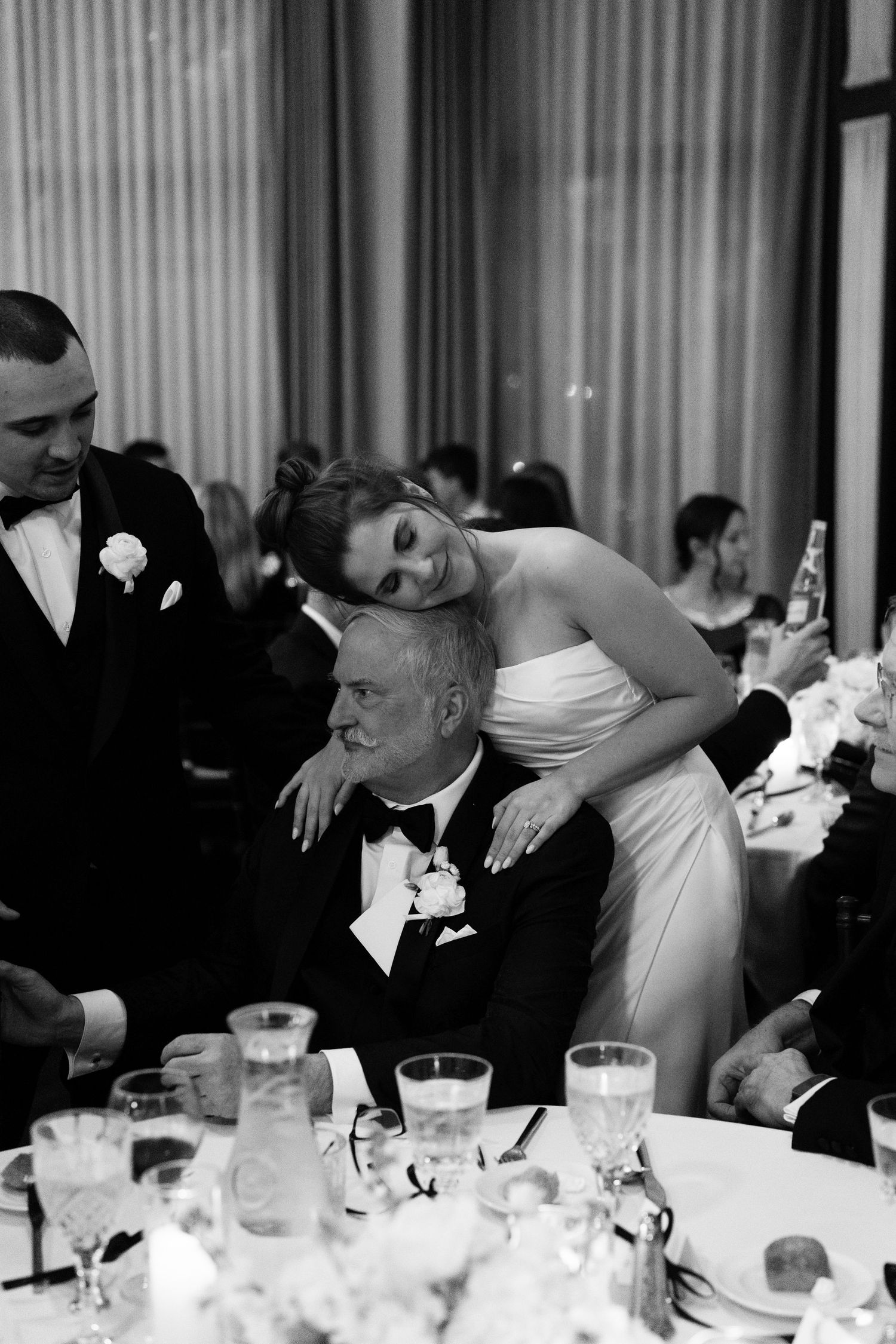 A bride leans over her father during a formal wedding reception dinner at an elegant banquet table.