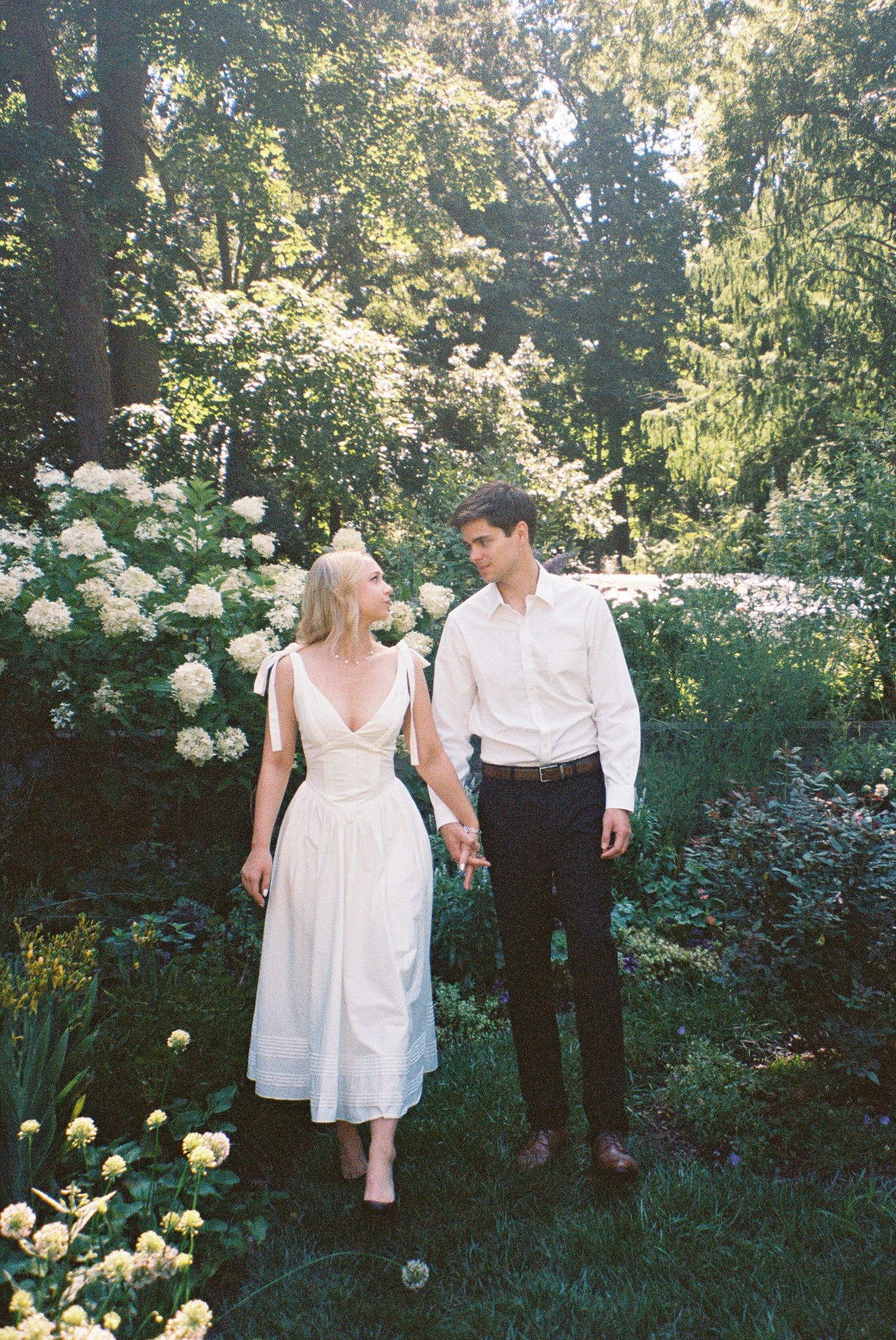 A couple in white attire holds hands while walking through a lush garden with blooming white roses in Newfields