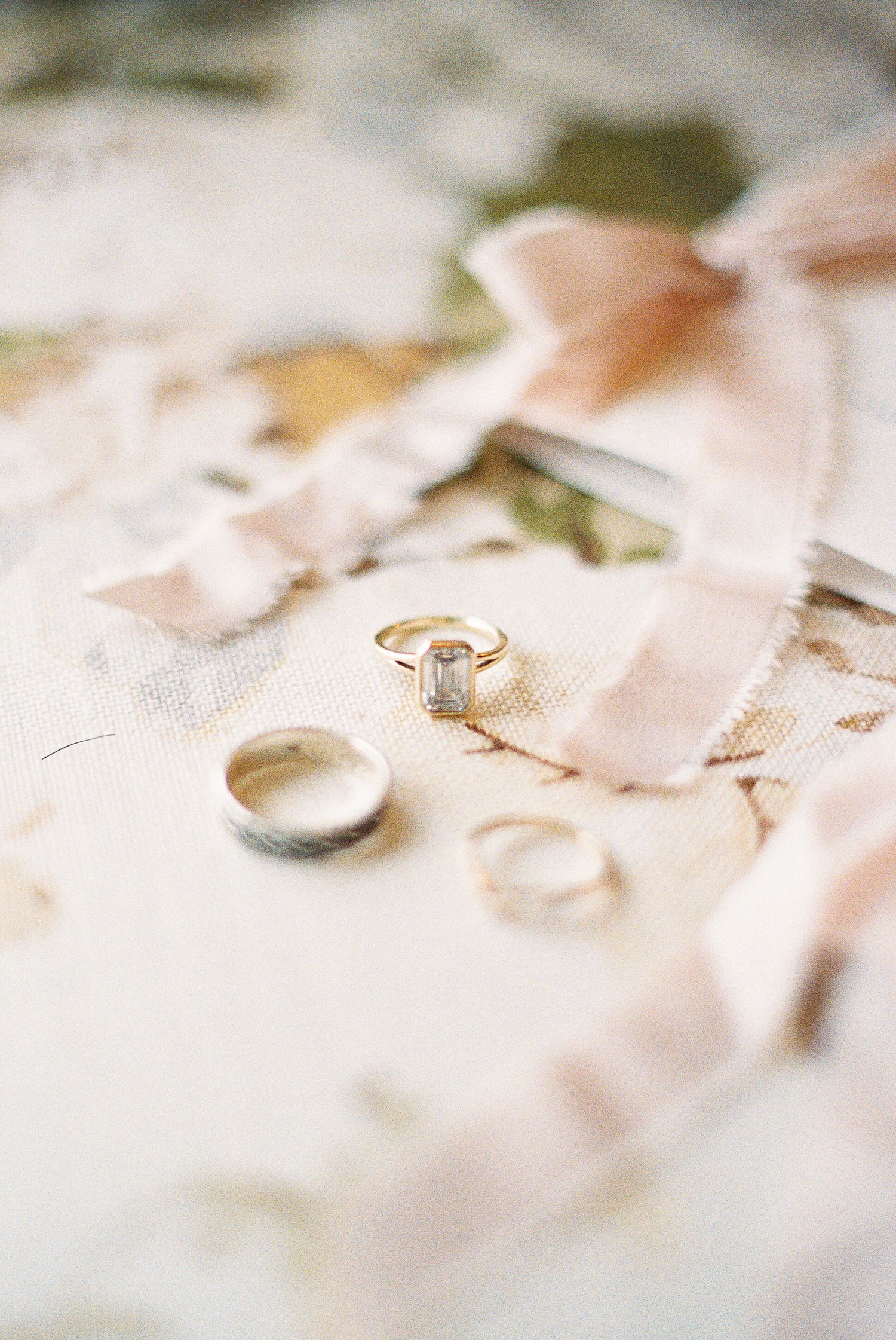 Gold engagement ring with emerald-cut diamond beside wedding bands on floral fabric with blush silk ribbon on vintage film
