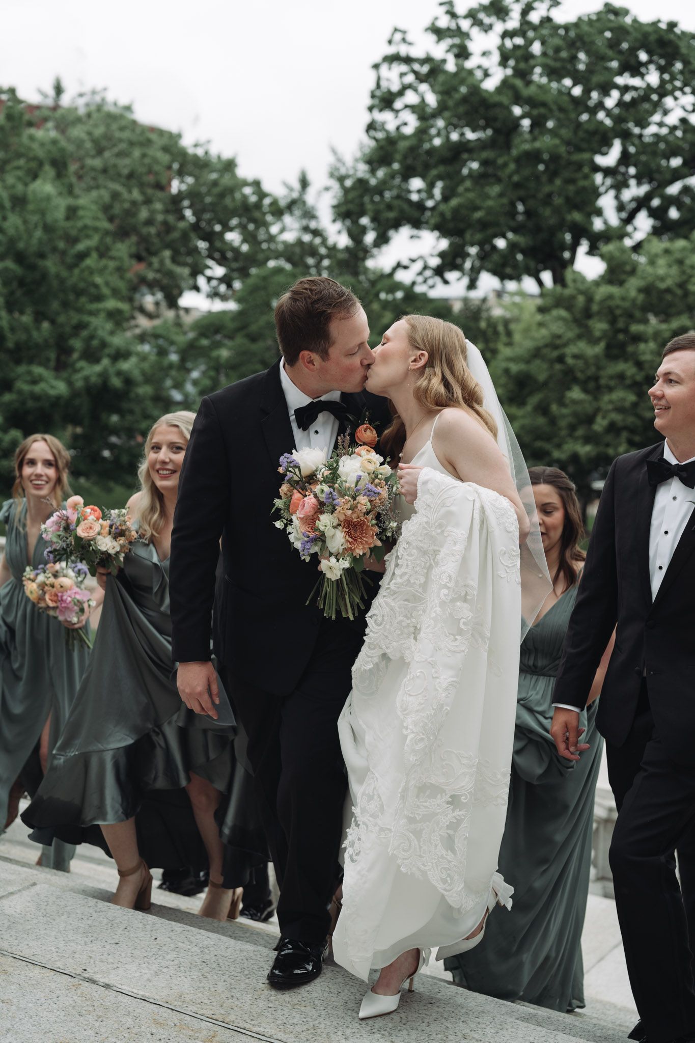 A tender kiss between newlyweds at an outdoor wedding celebration with bridesmaids in green dresses looking on.