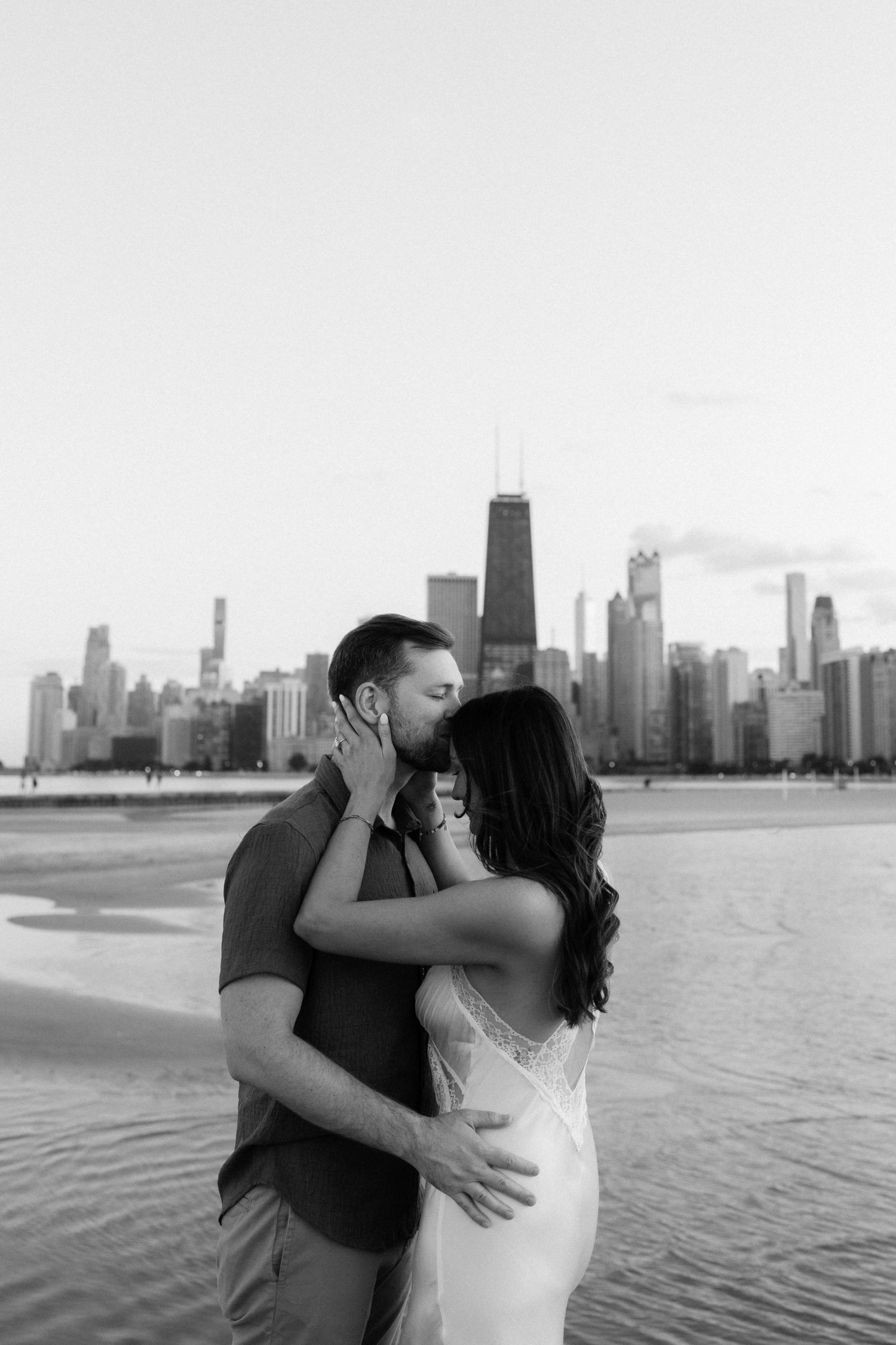 A romantic couple embraces by the Chicago waterfront with the iconic skyline and John Hancock building in the background.