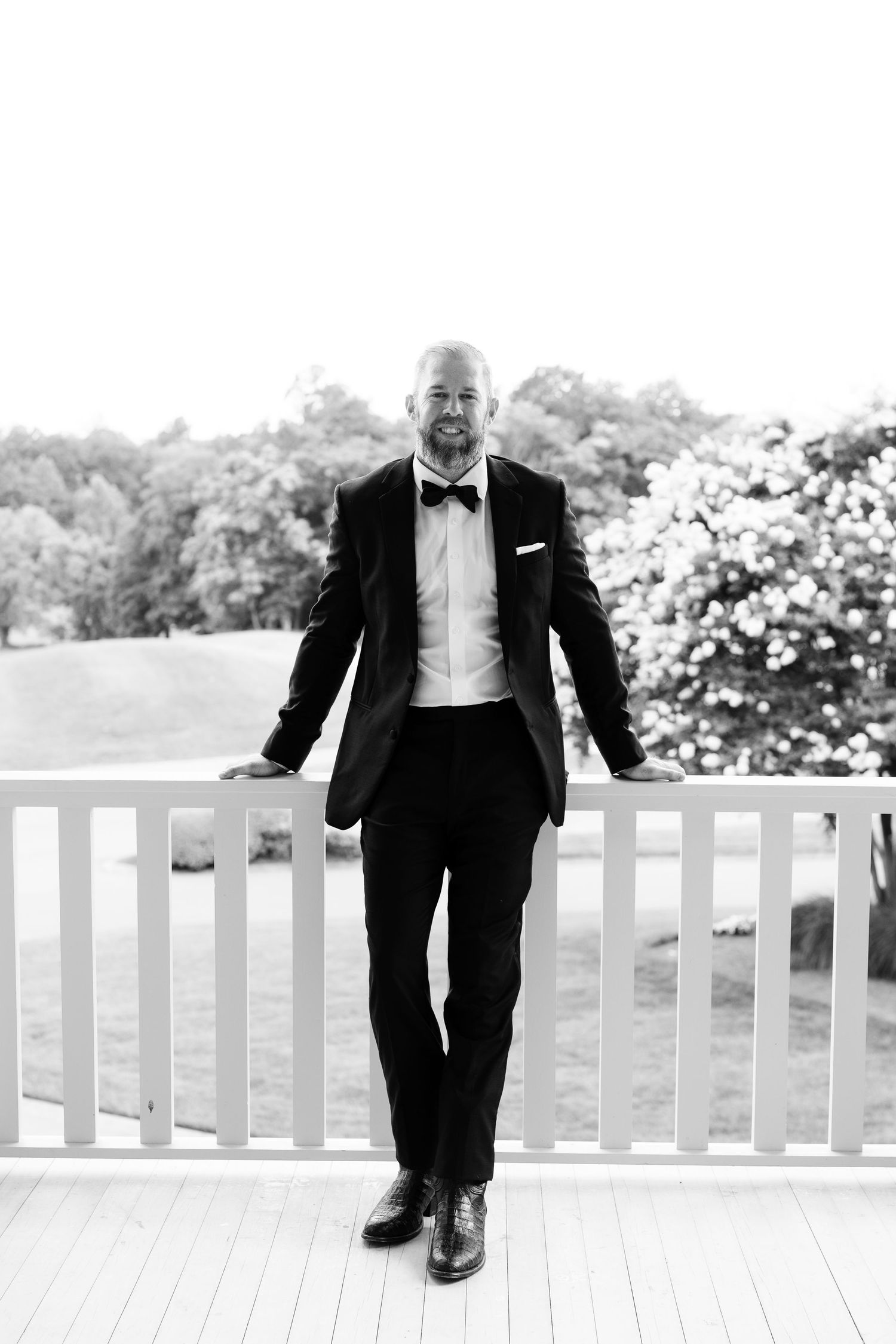 Groom relaxes casually on a white porch railing wearing formal black tie attire in Nashville, Tennessee