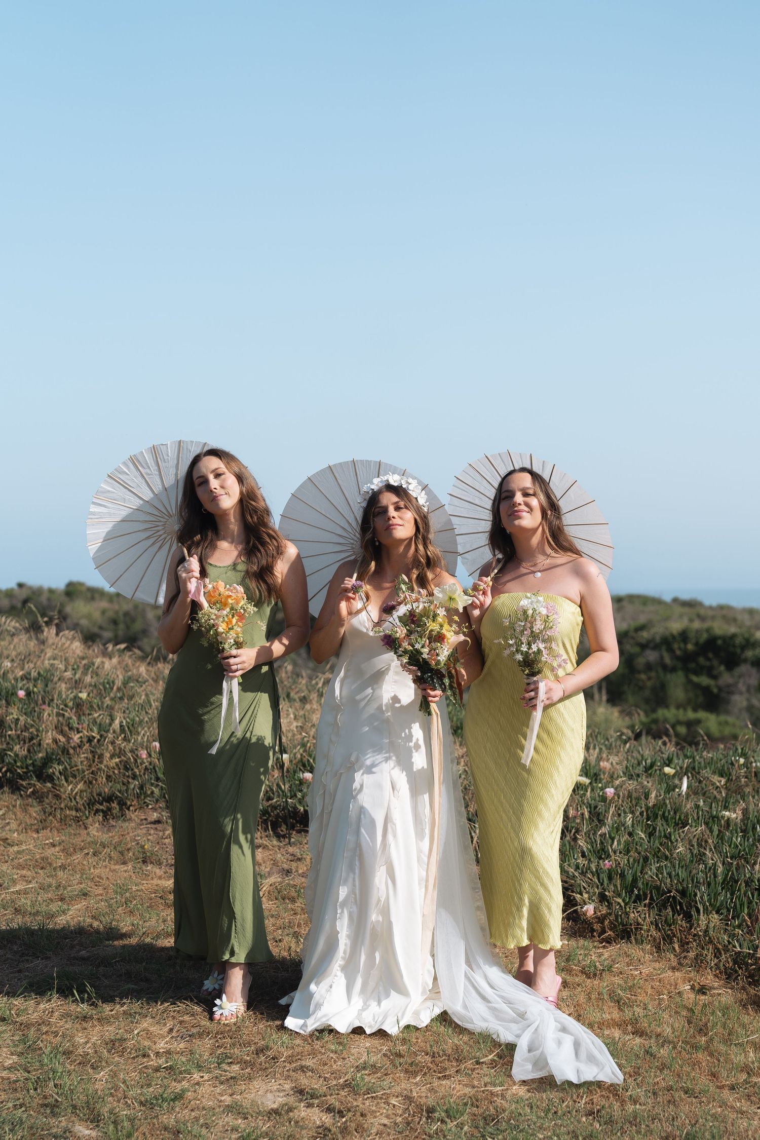 Three women in wedding attire pose with parasols on a grassy hillside during a sunny outdoor photoshoot in California