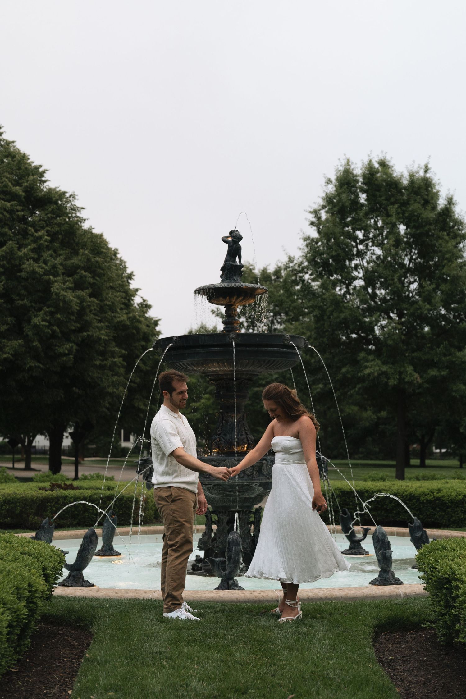 Couple holding hands in front of an ornate fountain with landscaped gardens and trees in the background in Carmel, Indiana