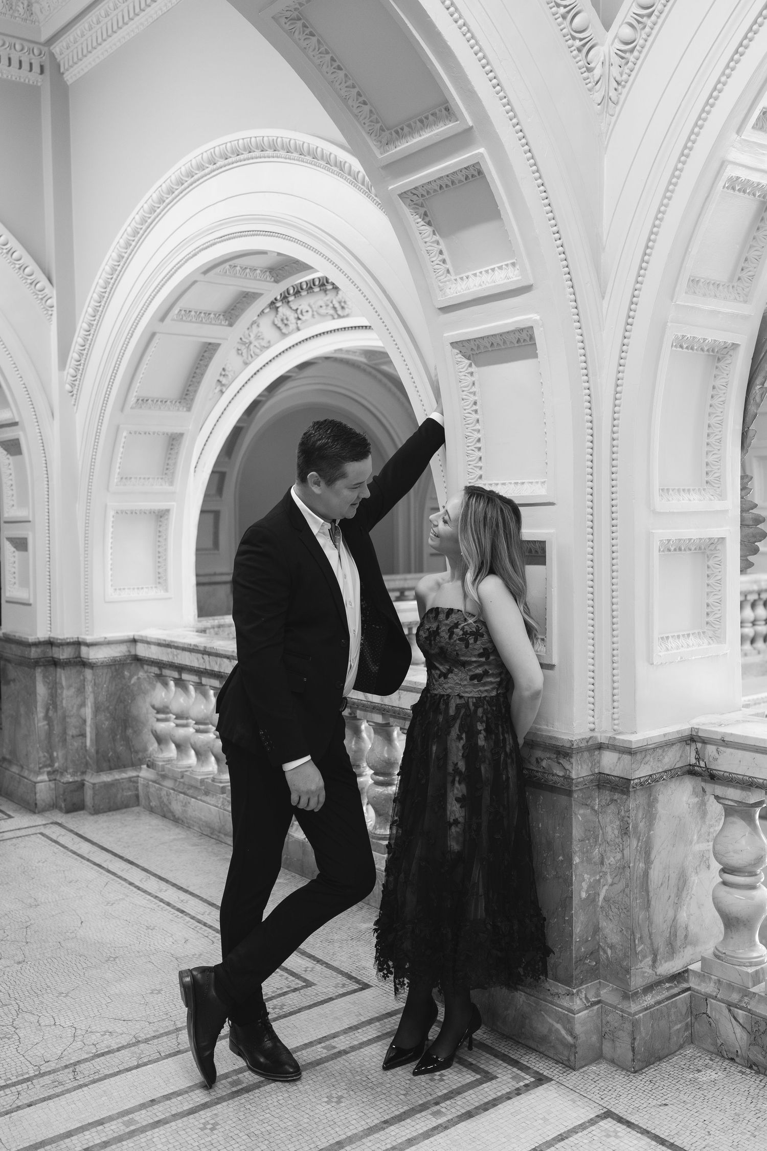Black and white photo of an elegant couple dancing in a grand arched hallway, dressed in formal attire.