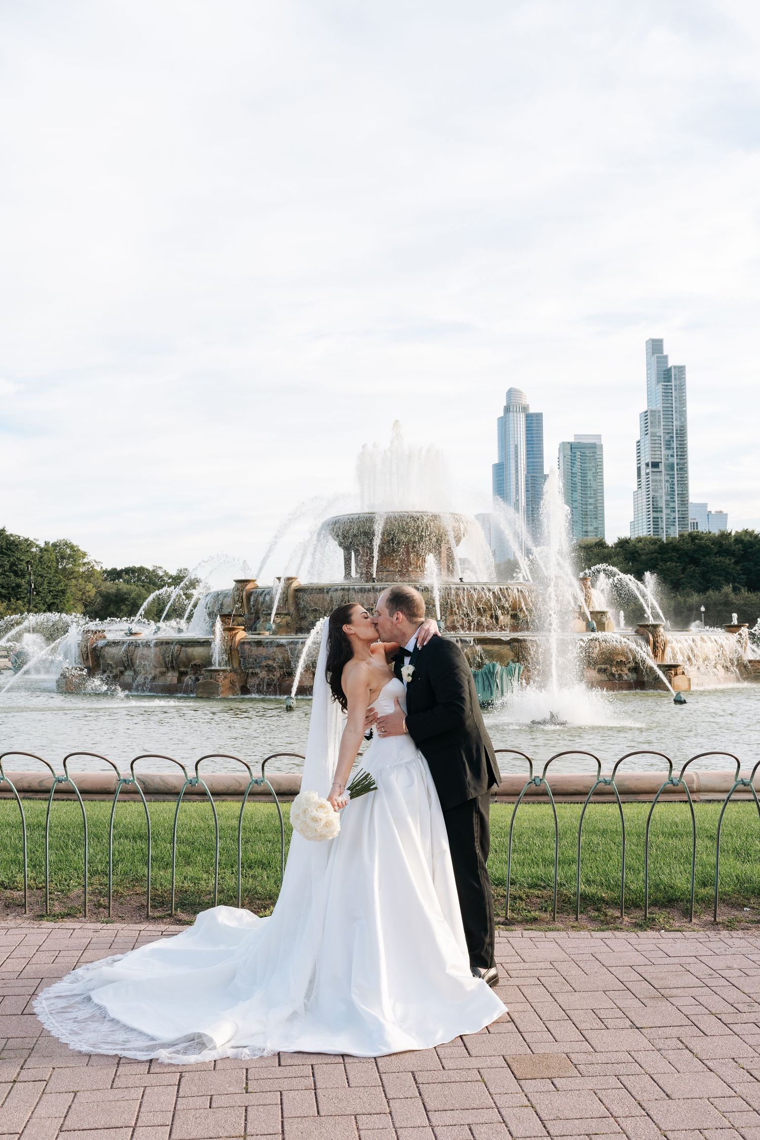 Bride in a white gown and groom in a black tuxedo kiss in front of Buckingham Fountain with the Chicago skyline behind them.
