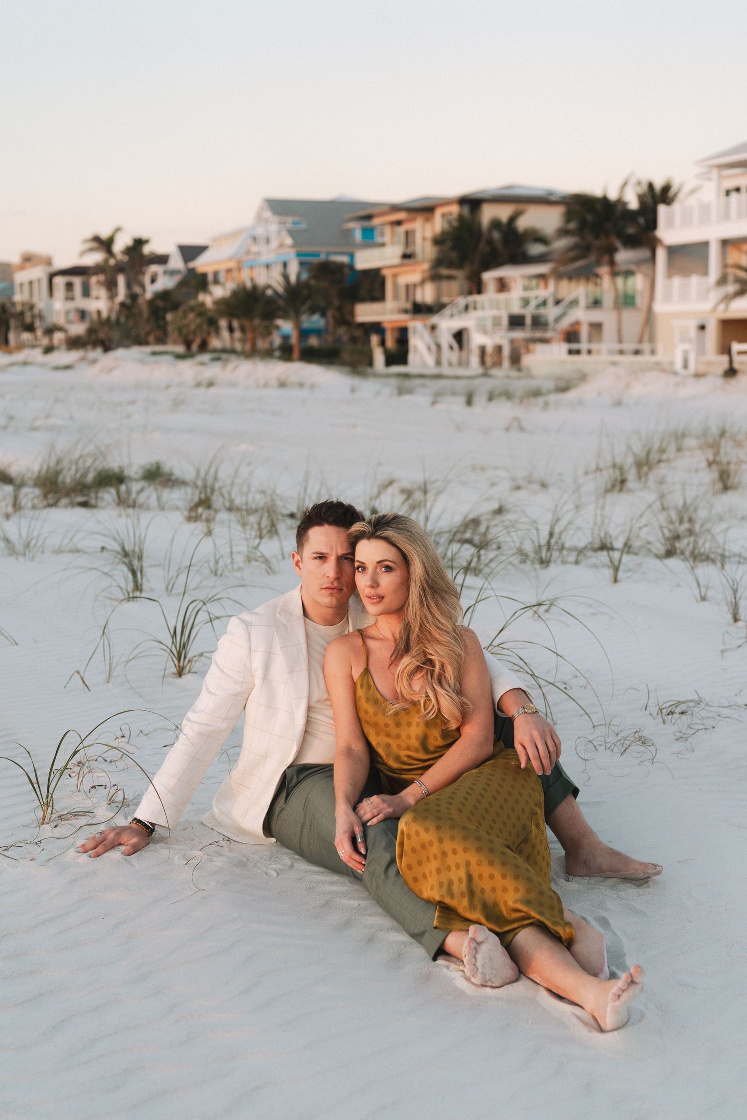 A couple sits together on white sand beach at sunset with beachfront homes and palm trees in Clearwater, Florida