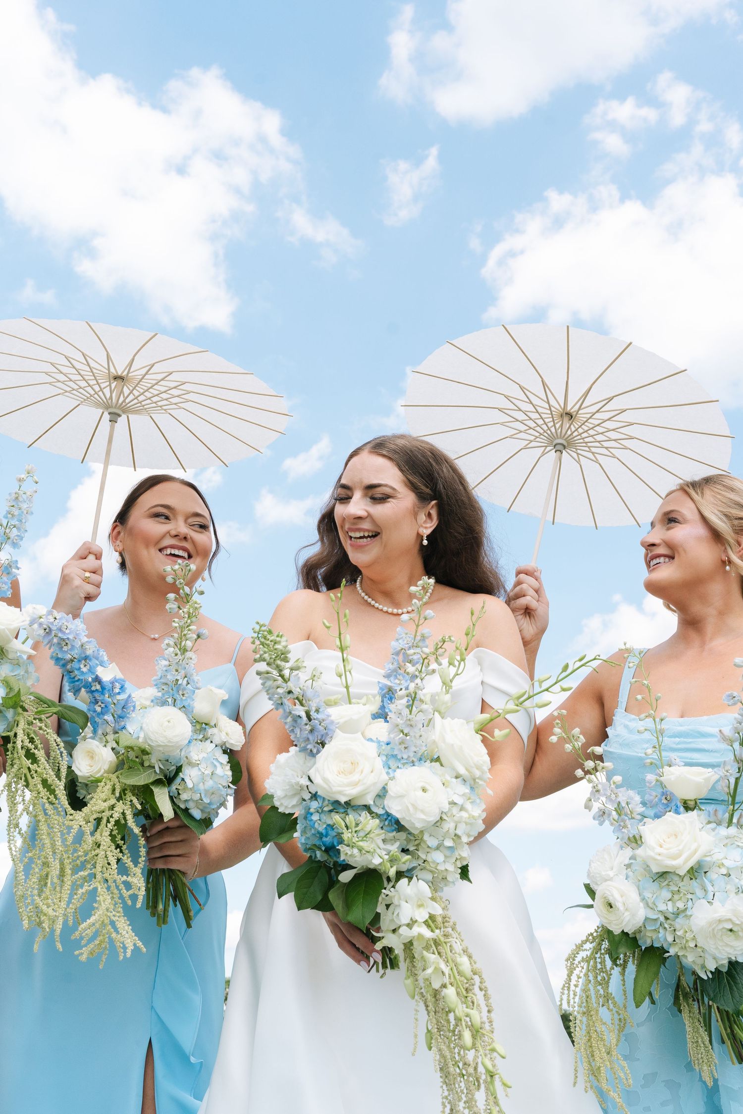 Bride in white gown with bridesmaids in blue dresses holding white bouquets and parasols under a blue sky in Illinois