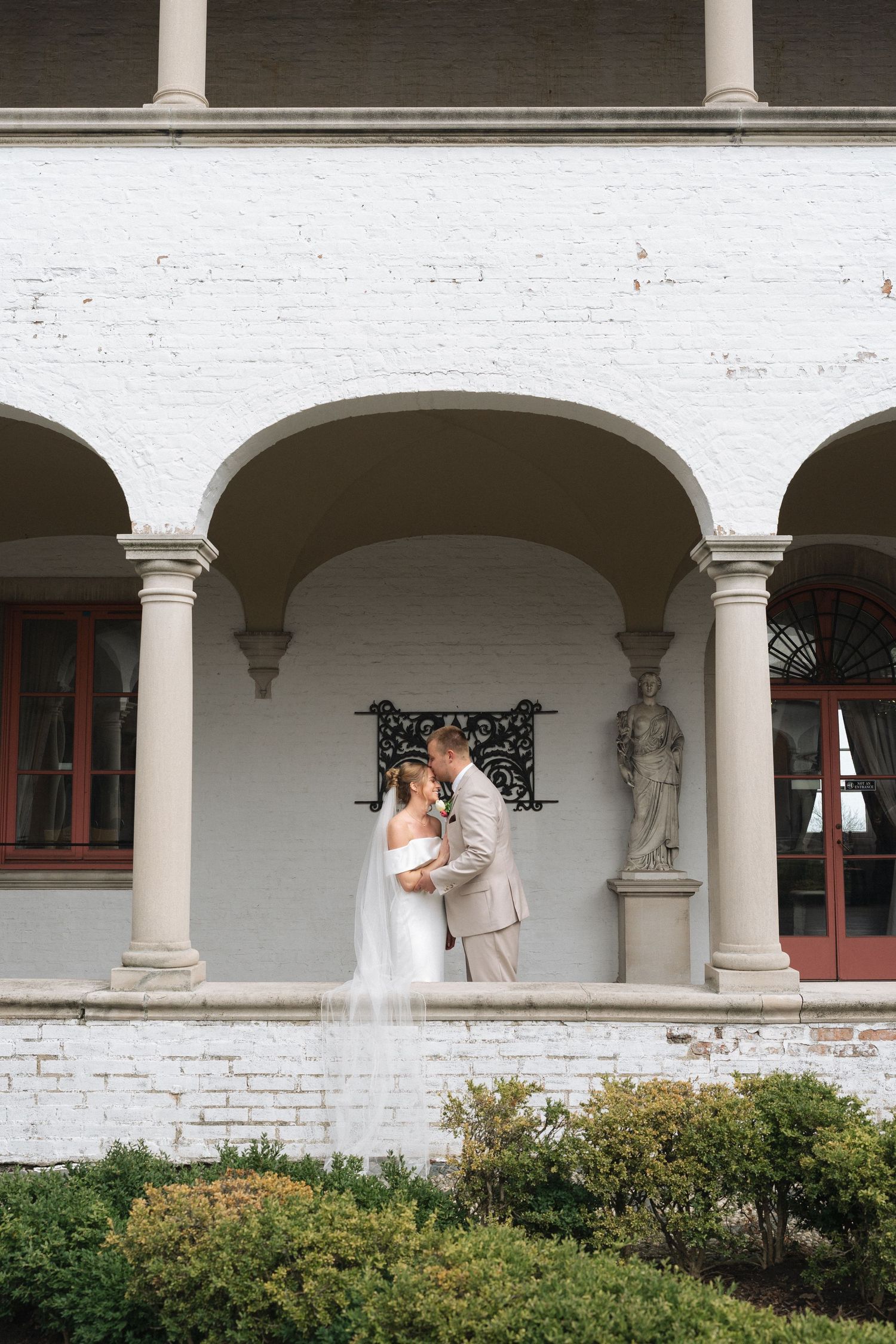 Bride and groom share a romantic kiss under elegant European arched colonnade of a white brick Italian villa with classical columns.