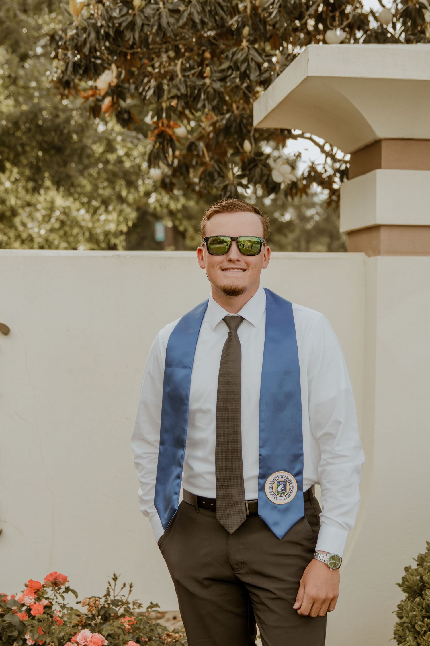 A graduate in sunglasses wearing a white dress shirt, blue graduation stole, and dark tie poses outdoors.