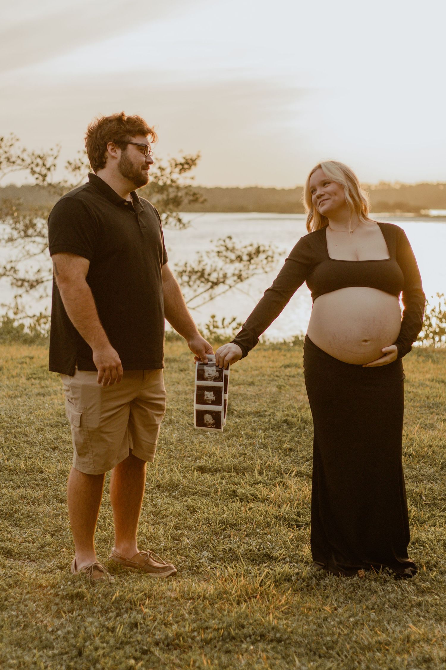 An expectant couple poses in a field at sunset holding ultrasound photos.